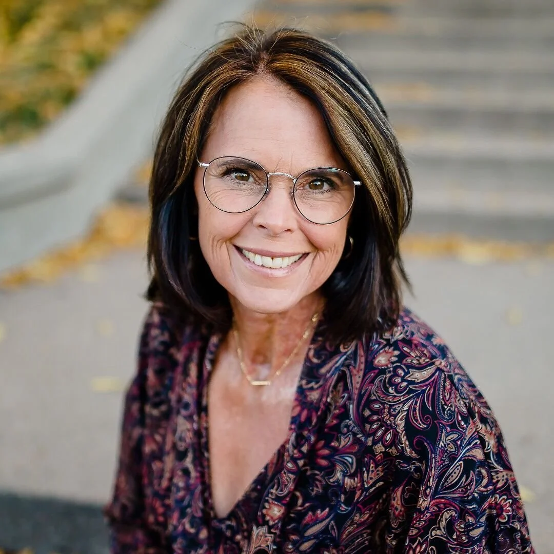 Smiling woman with glasses and shoulder-length dark hair with highlights, wearing a patterned blouse, outdoors with steps and fallen leaves in the background.