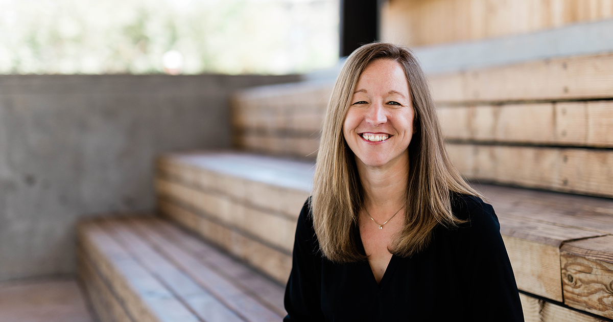 Smiling woman with long blond hair wearing a black top, sitting in front of wooden bleacher stairs.