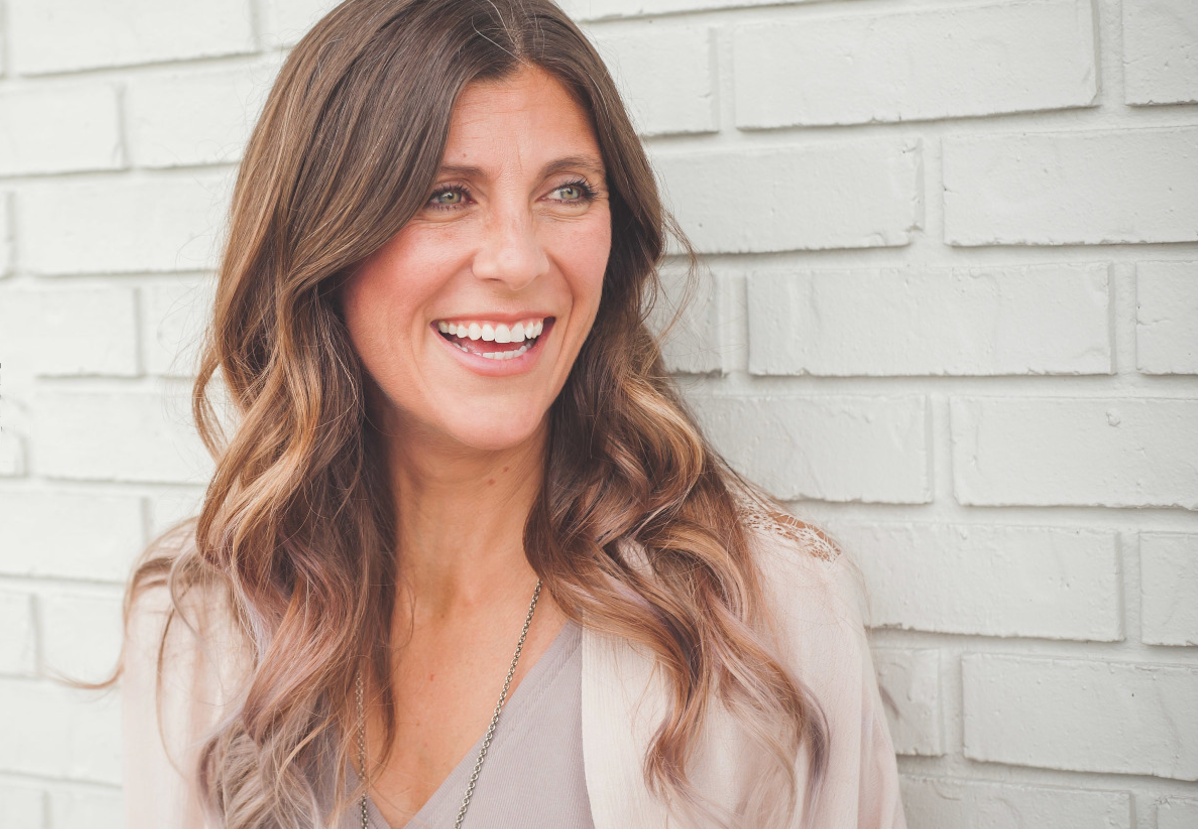 A woman with long, wavy brown hair smiles while standing against a white brick wall.