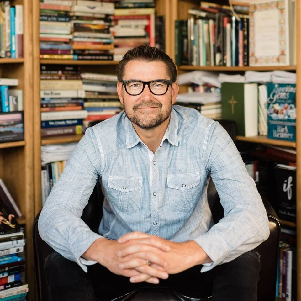 A man wearing glasses and a light blue button-up shirt sits in front of a bookshelf filled with books, smiling at the camera.