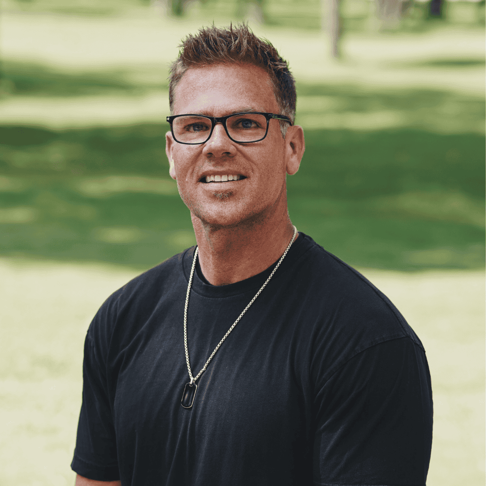 A man with glasses and short brown hair outdoors, wearing a black t-shirt and a necklace, smiling at the camera.