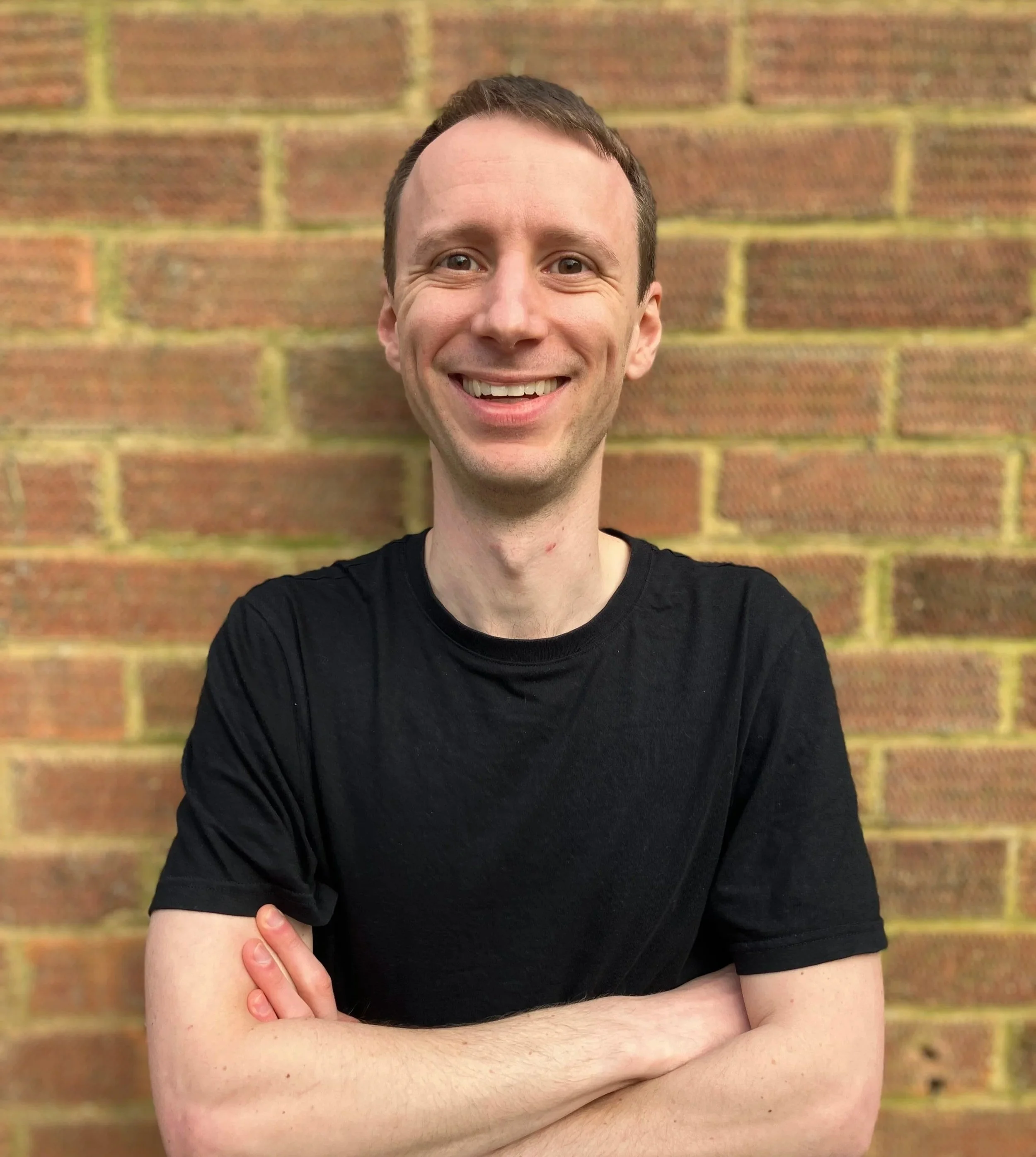A young man with short brown hair, smiling, wearing a black t-shirt, standing in front of a red brick wall.