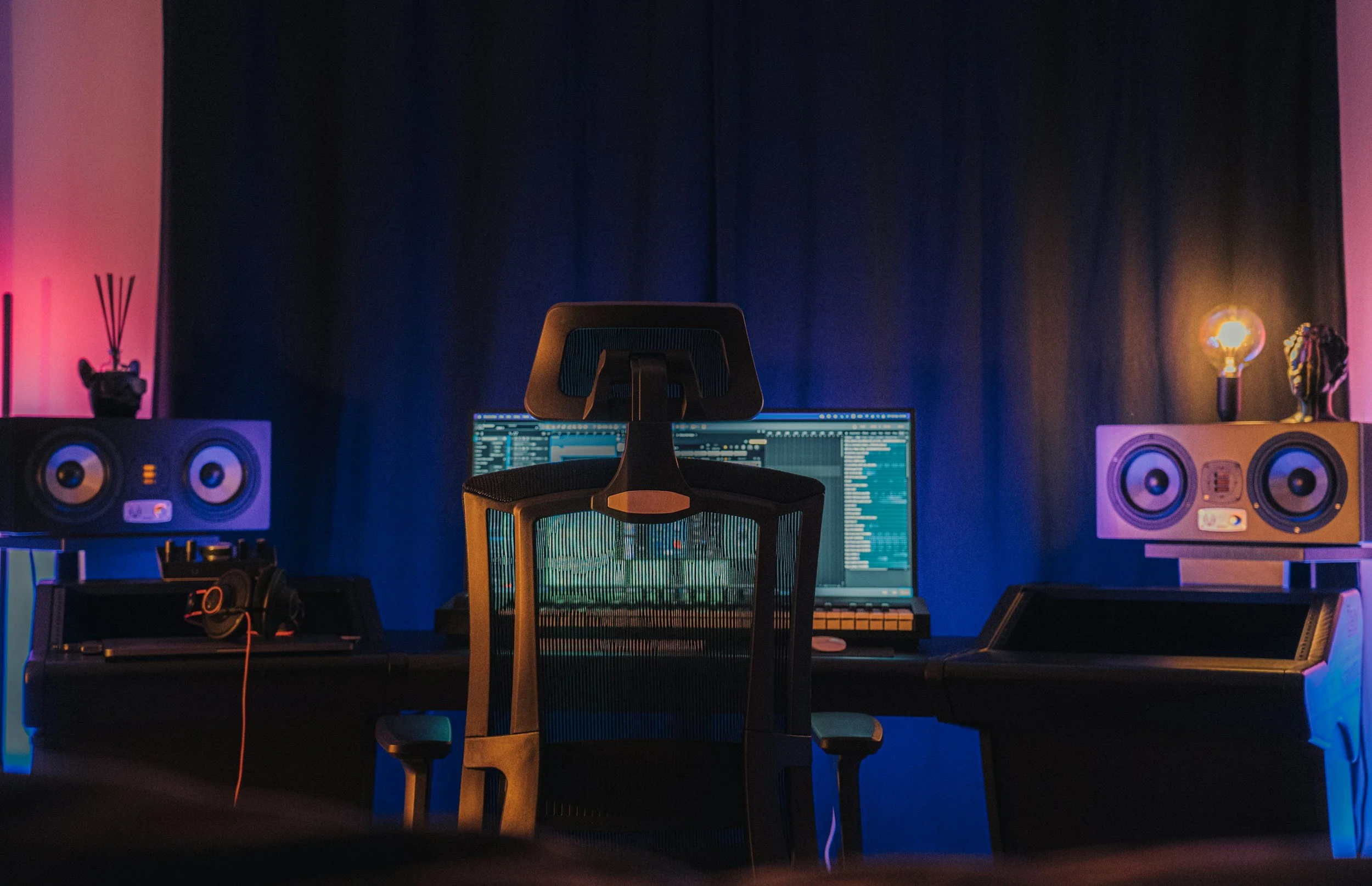 A music production workspace featuring a computer monitor displaying audio editing software, studio monitors on either side, a professional ergonomic chair, and various electronic equipment in a room with purple and orange lighting.