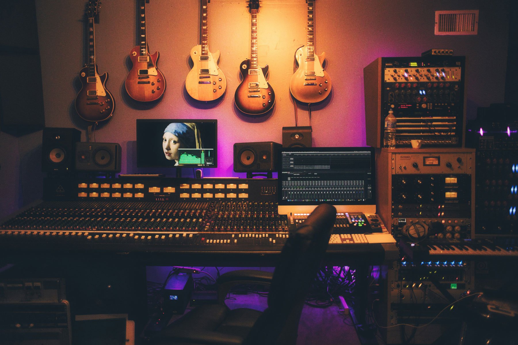 A music production studio featuring five electric guitars mounted on a wall above a mixing console and computer screens displaying audio editing software. The studio is illuminated with purple and warm lighting.