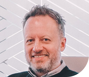 Close-up of a smiling man with short gray hair and beard, wearing a black shirt, in front of a modern, geometric white background.