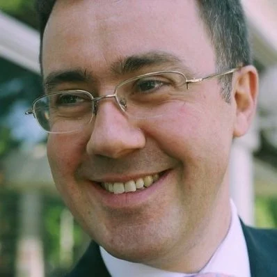 Close-up of a smiling man wearing glasses and a suit outside.