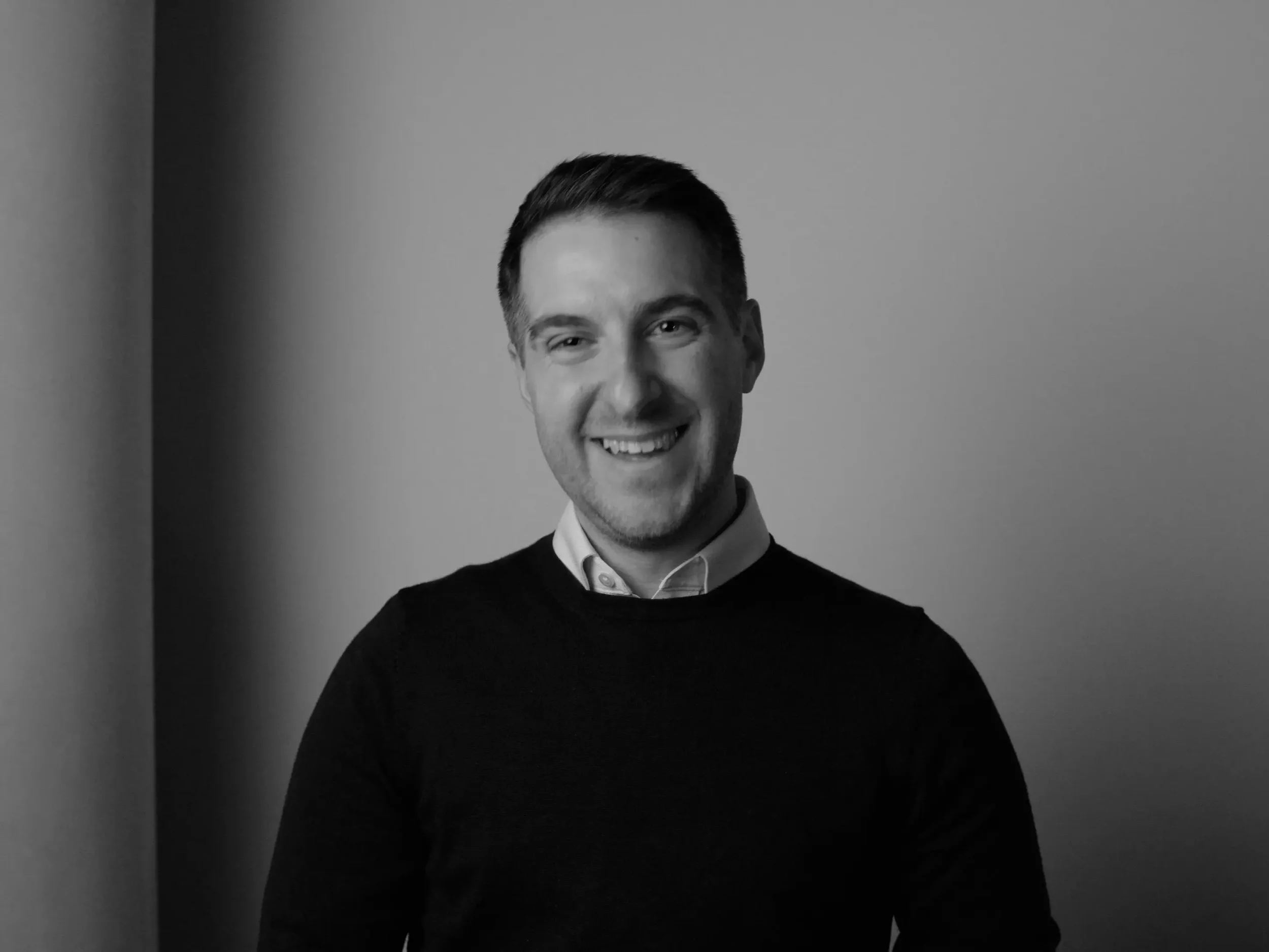 A black-and-white portrait of a smiling man with short dark hair, wearing a collared shirt and a dark sweater, positioned against a simple gradient background.
