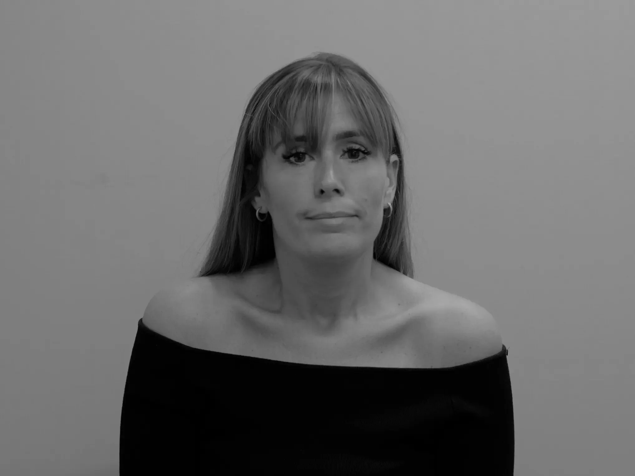 Black-and-white portrait of a woman with long hair and bangs, wearing a black off-the-shoulder top, looking directly at the camera against a plain background.