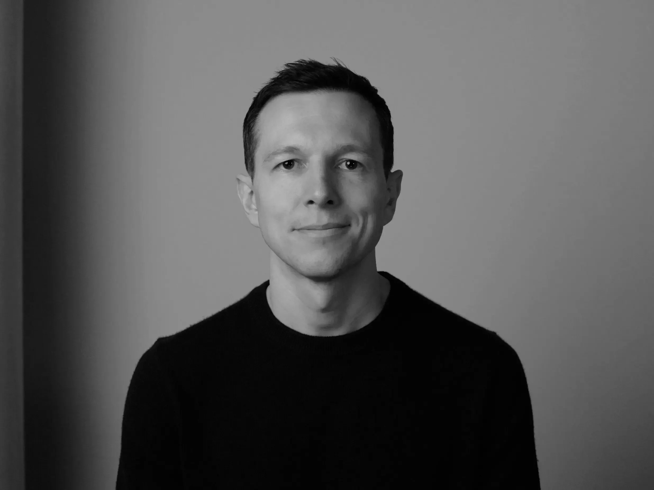 Black and white portrait of a young man with short dark hair, wearing a black crew neck shirt, standing against a plain background.