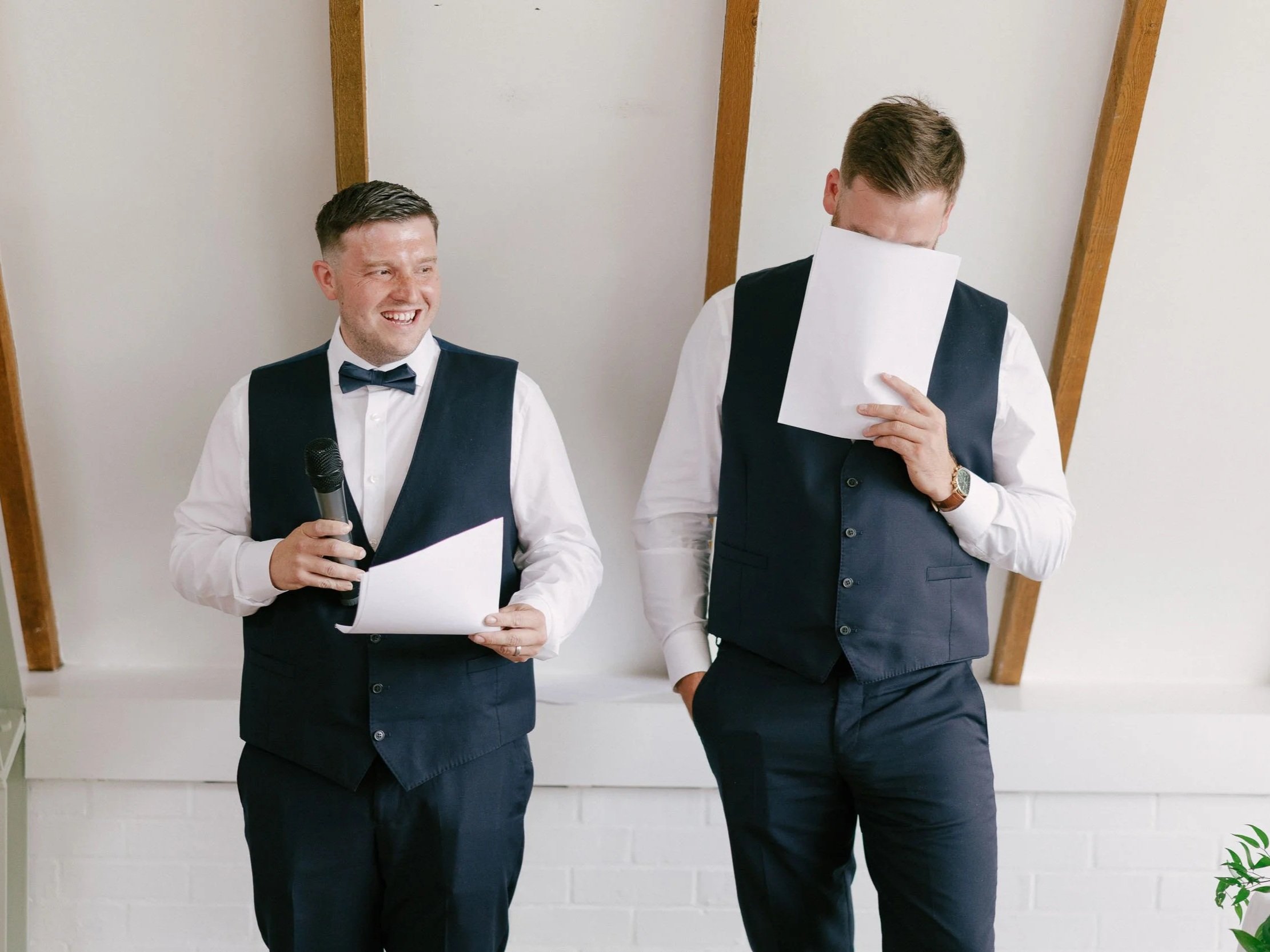Two men dressed in formal attire, one holding a microphone and paper, the other covering his face with a paper, standing against a white wall with wooden beams.