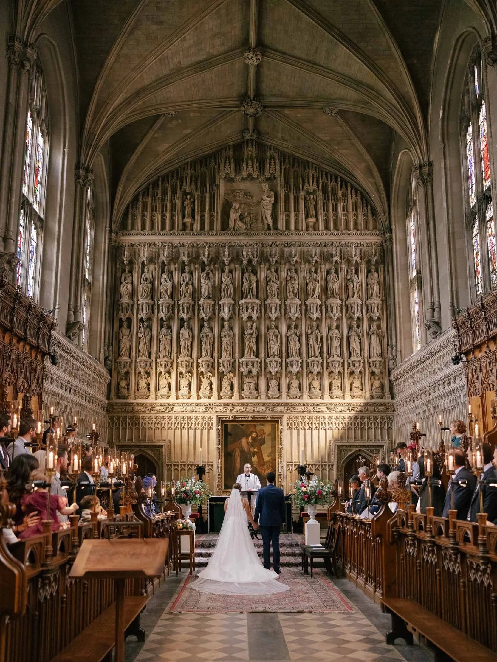 A wedding ceremony taking place inside a large, ornate church with high vaulted ceilings and stained glass windows. The bride and groom stand at the altar, facing a priest, with guests seated on both sides of the aisle.