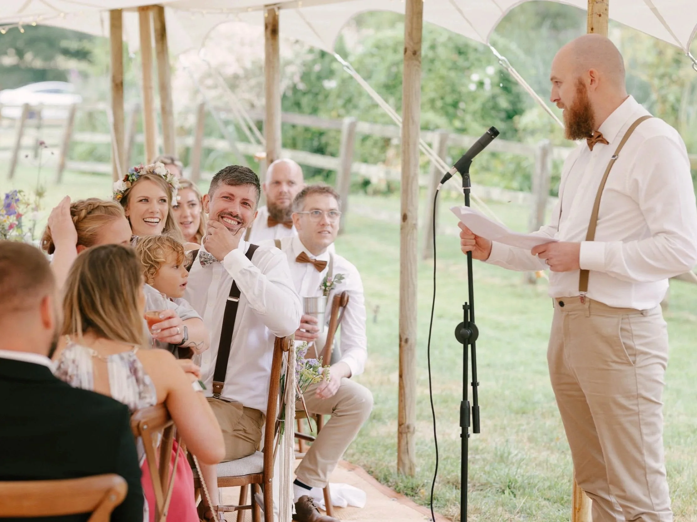 Man giving wedding vows or speech at outdoor wedding reception under a canopy, with seated guests, including children and a bride with floral crown, smiling and listening.