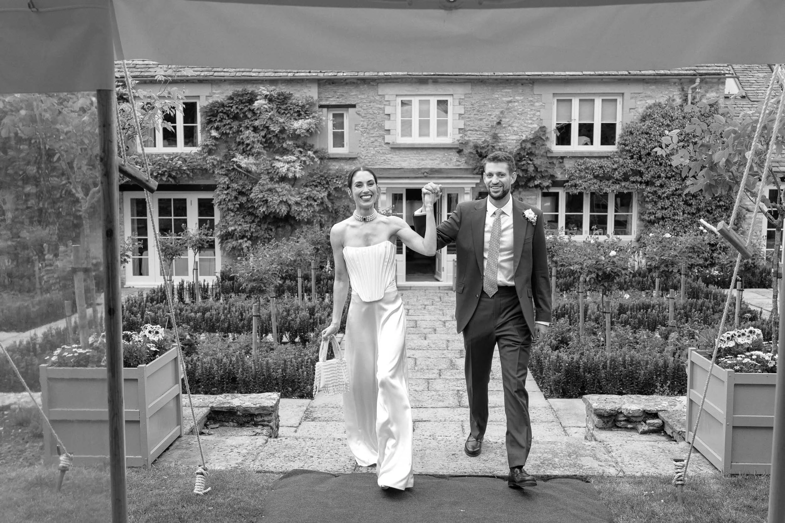 A bride and groom walking hand in hand outdoors in front of a house, smiling and celebrating their wedding day.