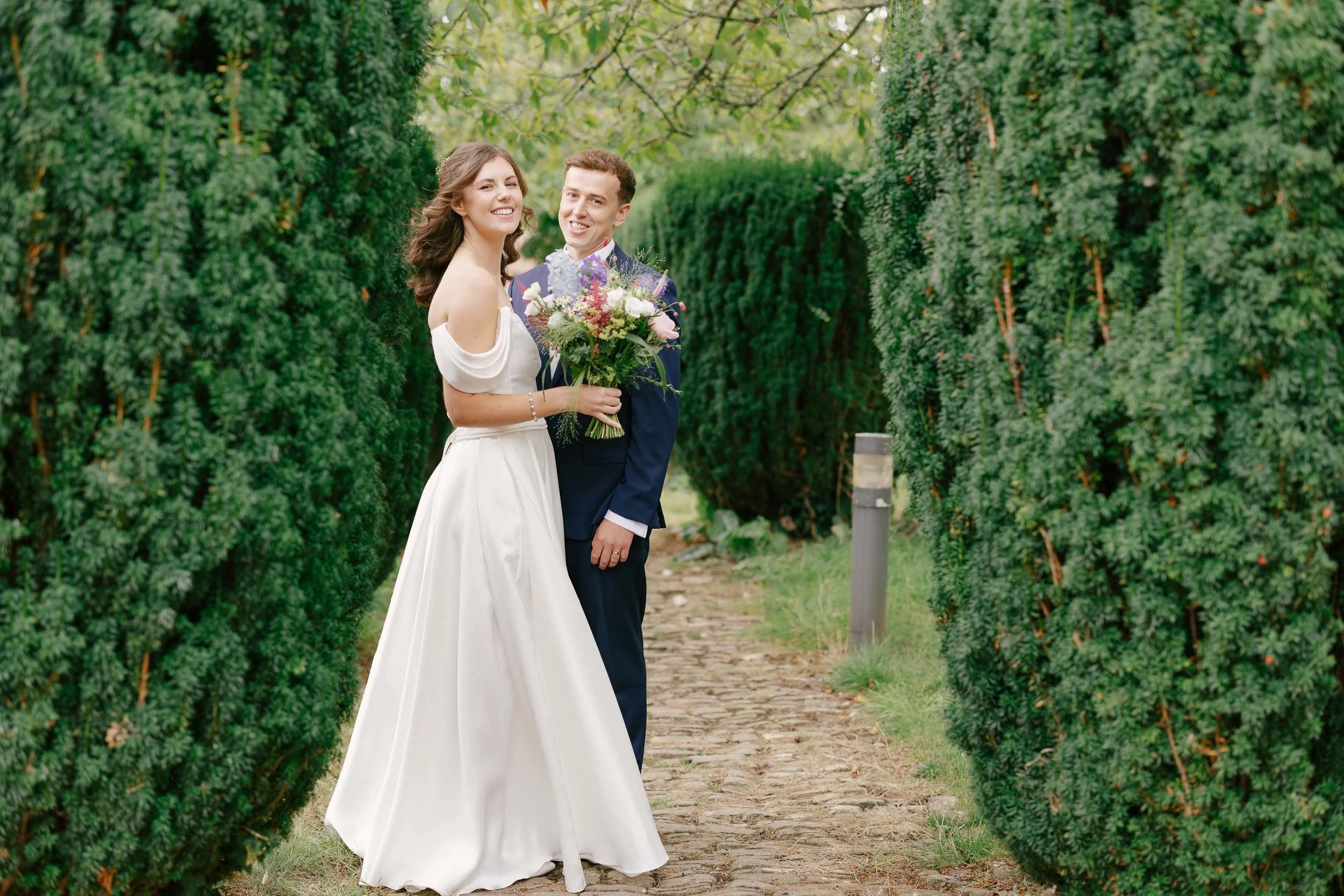 A bride in a white wedding dress holding a bouquet of flowers and a groom in a navy suit standing on a stone path between tall, green hedges during a wedding photoshoot.