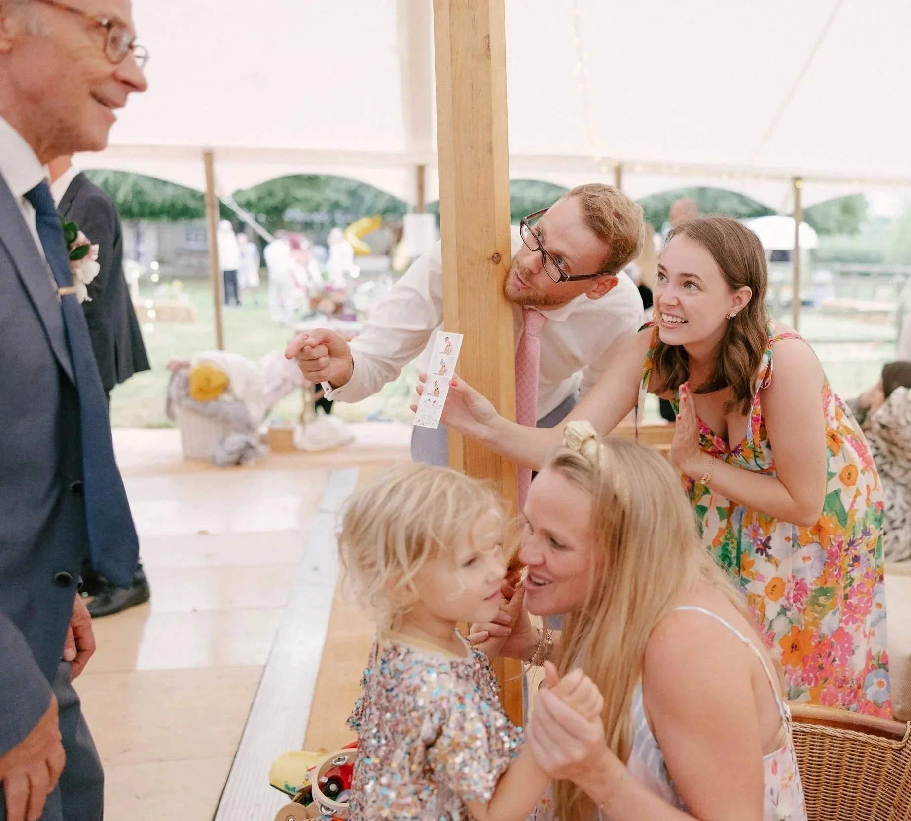 People at a celebration, including a woman with a child, a man in glasses, and a smiling woman in a floral dress, all interacting happily under a tent.
