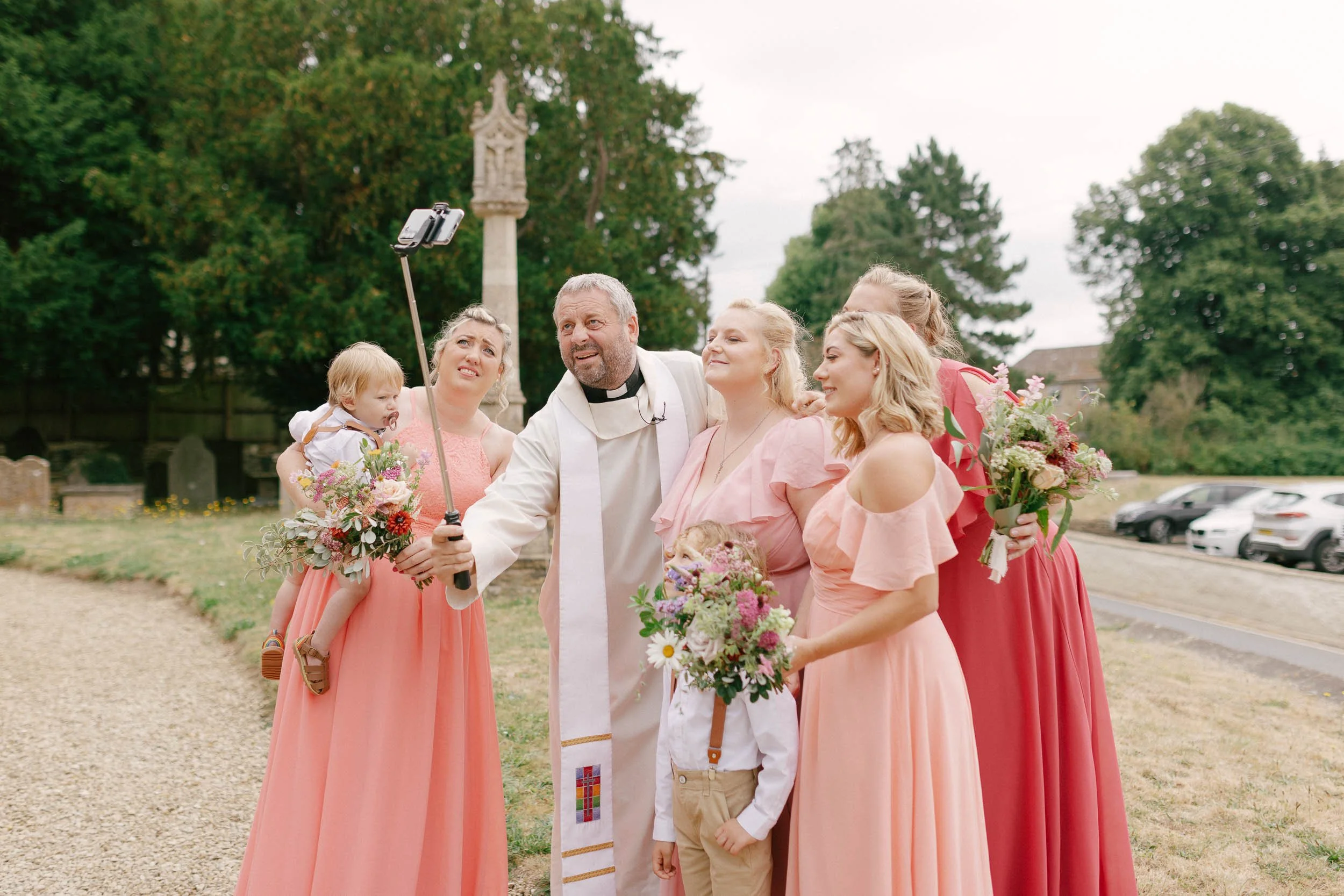 A group of people, including a priest and women in pink dresses, taking a selfie outside during a wedding celebration, with some holding floral bouquets.