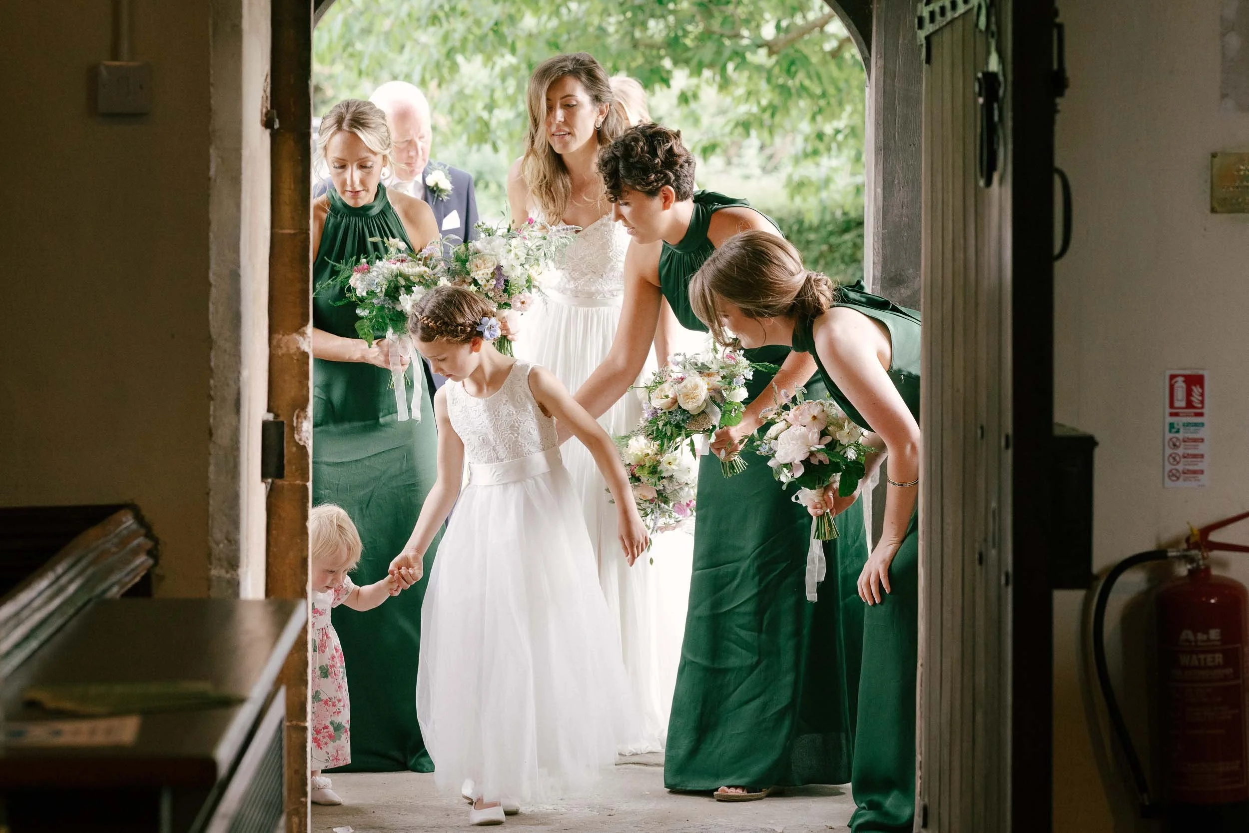 A bride in a white wedding dress and bridesmaids in green dresses help a young girl in a white dress and a toddler in a floral dress walk through a doorway during a wedding ceremony.