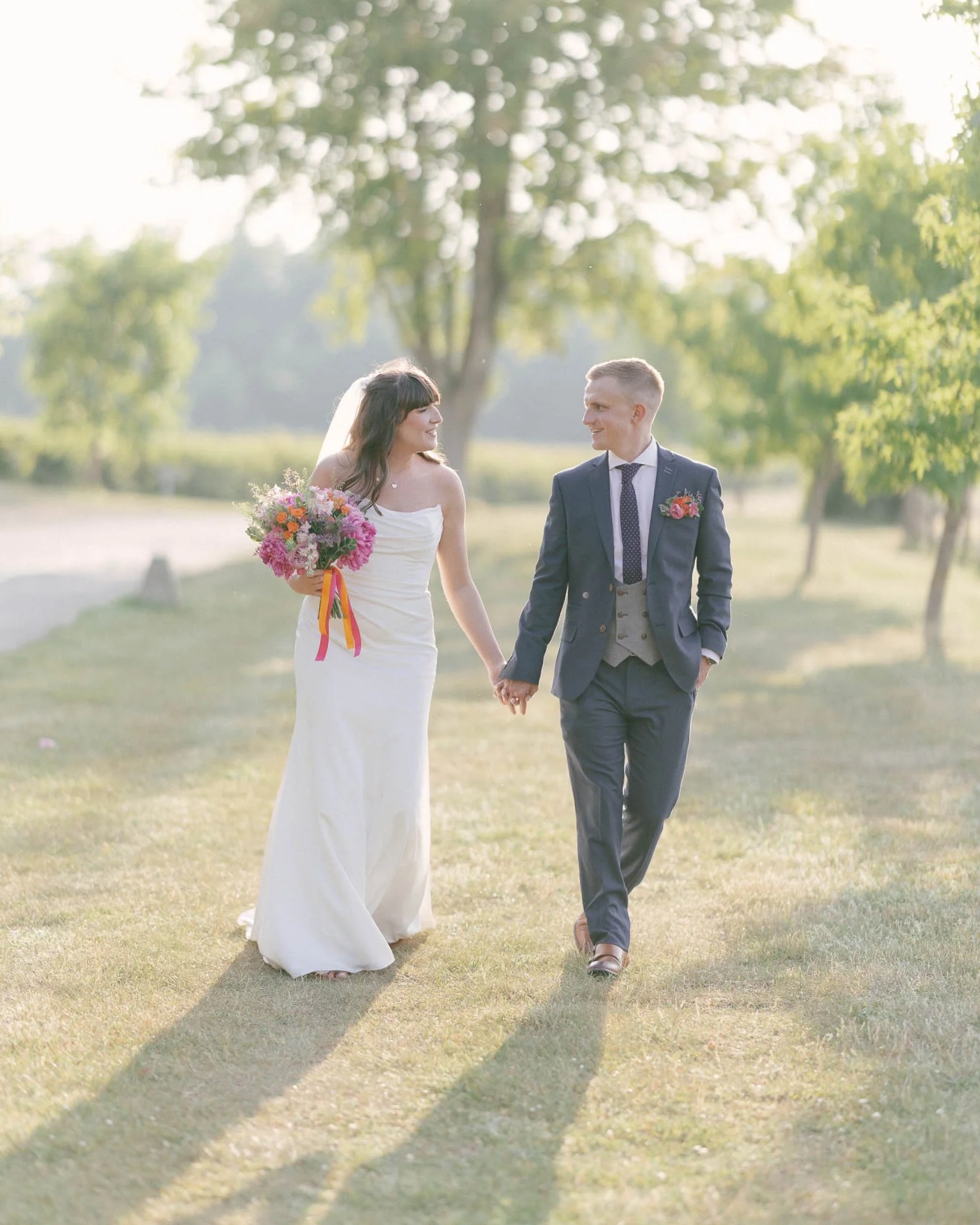 A bride and groom walking hand-in-hand outdoors at sunset, with lush green trees in the background. The bride is wearing a white wedding dress and holding a bouquet of colorful flowers, while the groom is dressed in a navy suit with a light gray vest and tie. They are smiling and looking at each other.