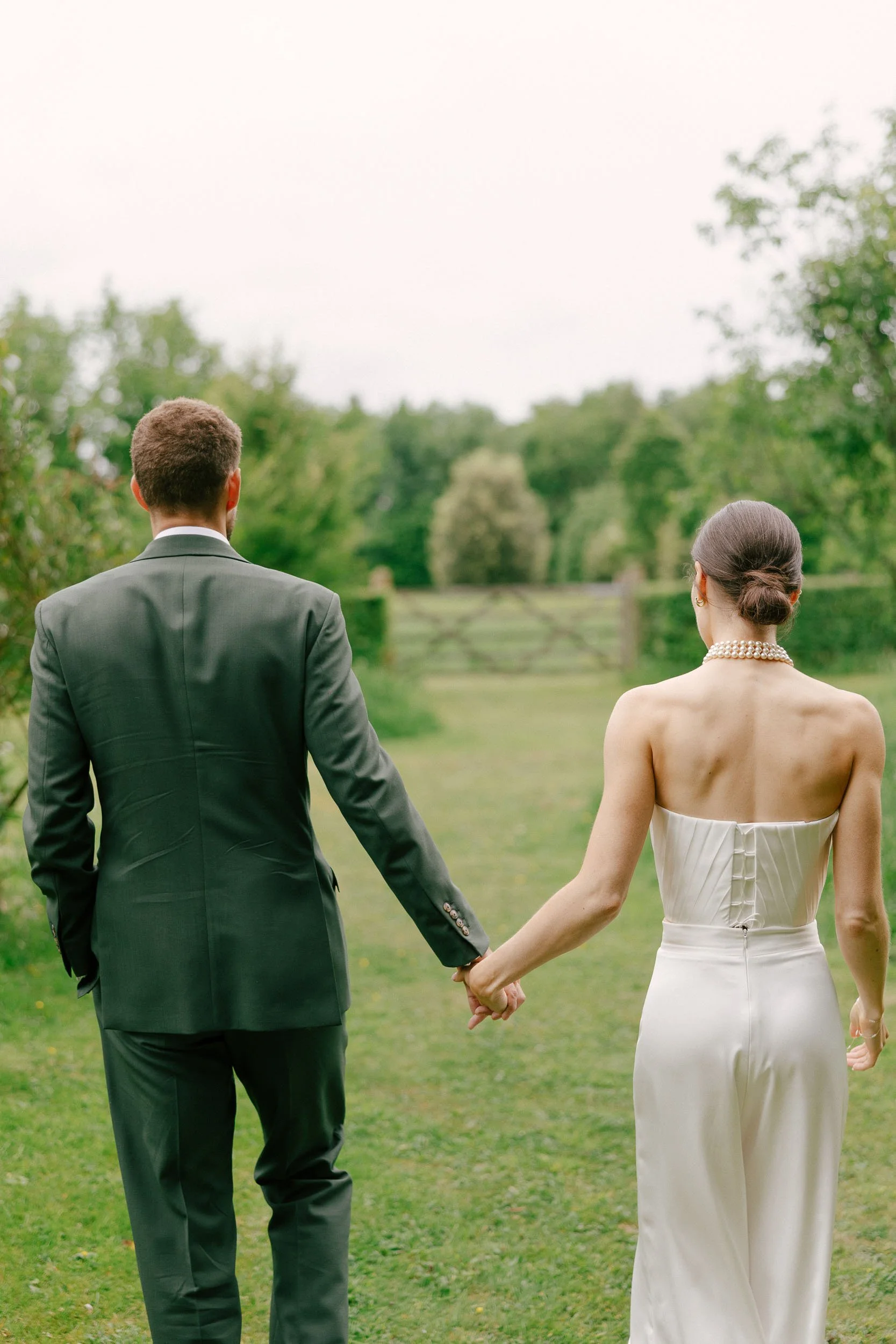 A couple dressed in wedding attire holding hands outdoors, walking away from camera on a grassy path surrounded by trees.