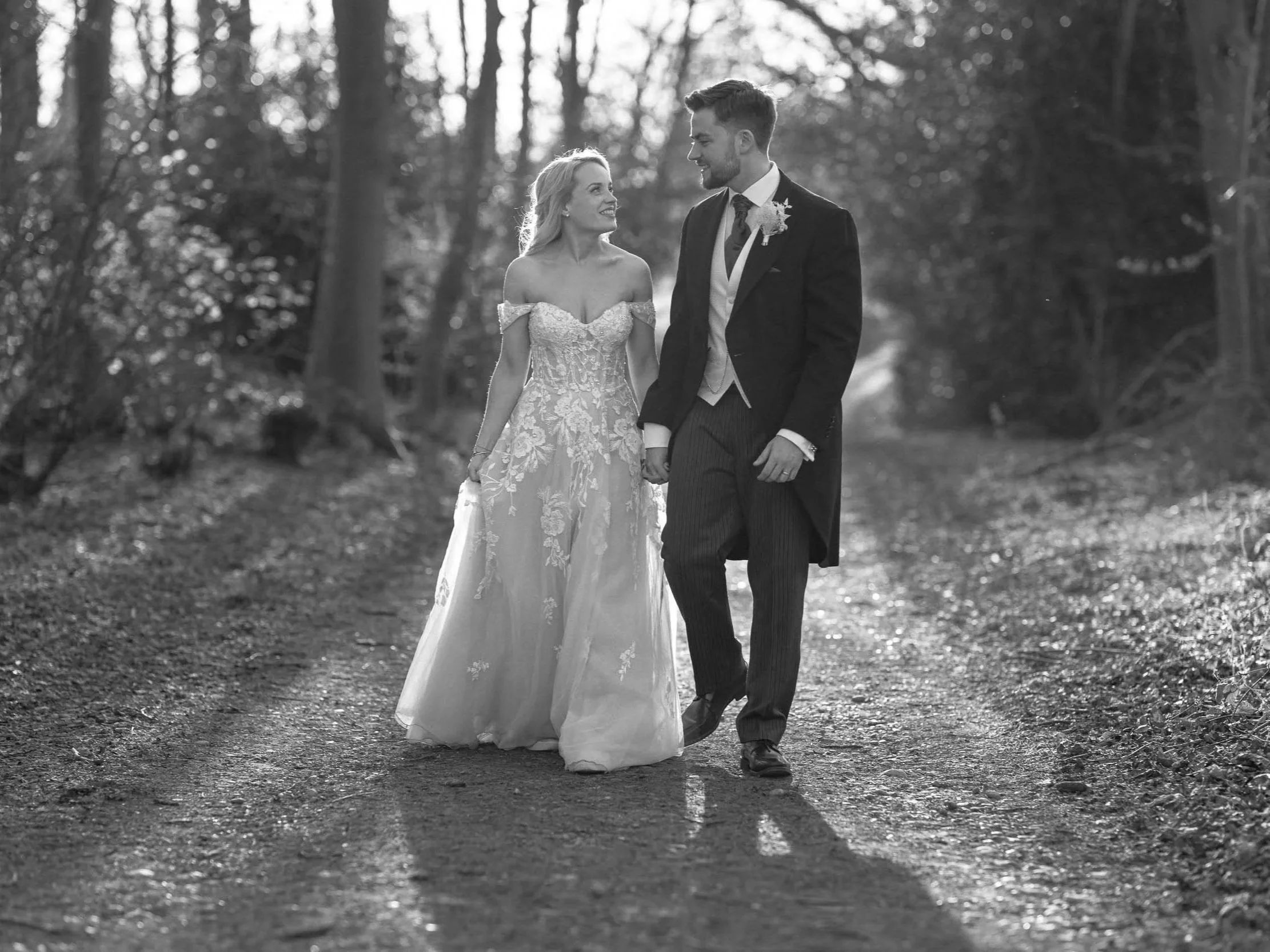 A bride and groom walking hand in hand on a wooded trail. The bride is in a lace wedding dress, and the groom is in a formal suit with a waistcoat and tie. The photo is in black and white.