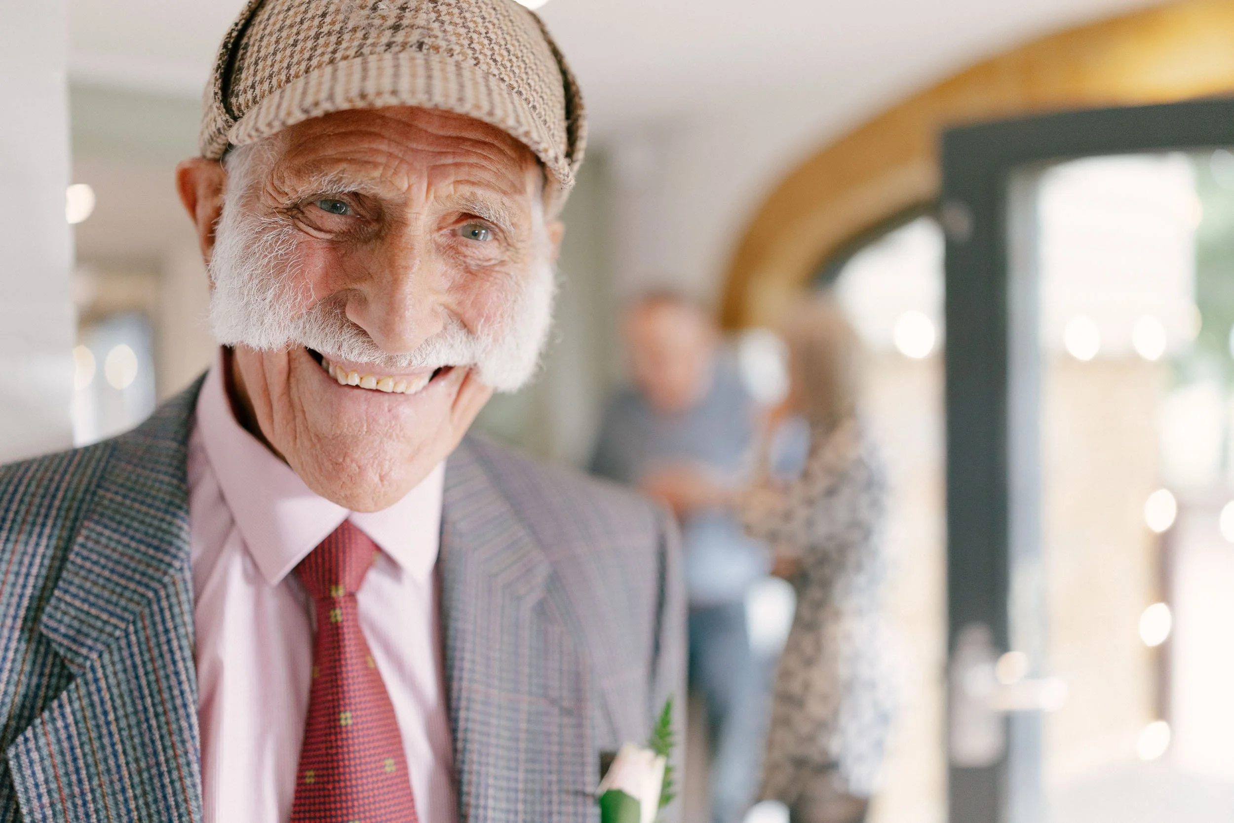 An elderly man with a white beard and mustache smiling, wearing a plaid jacket, pink shirt, red tie, and a checkered cap, at a social gathering.