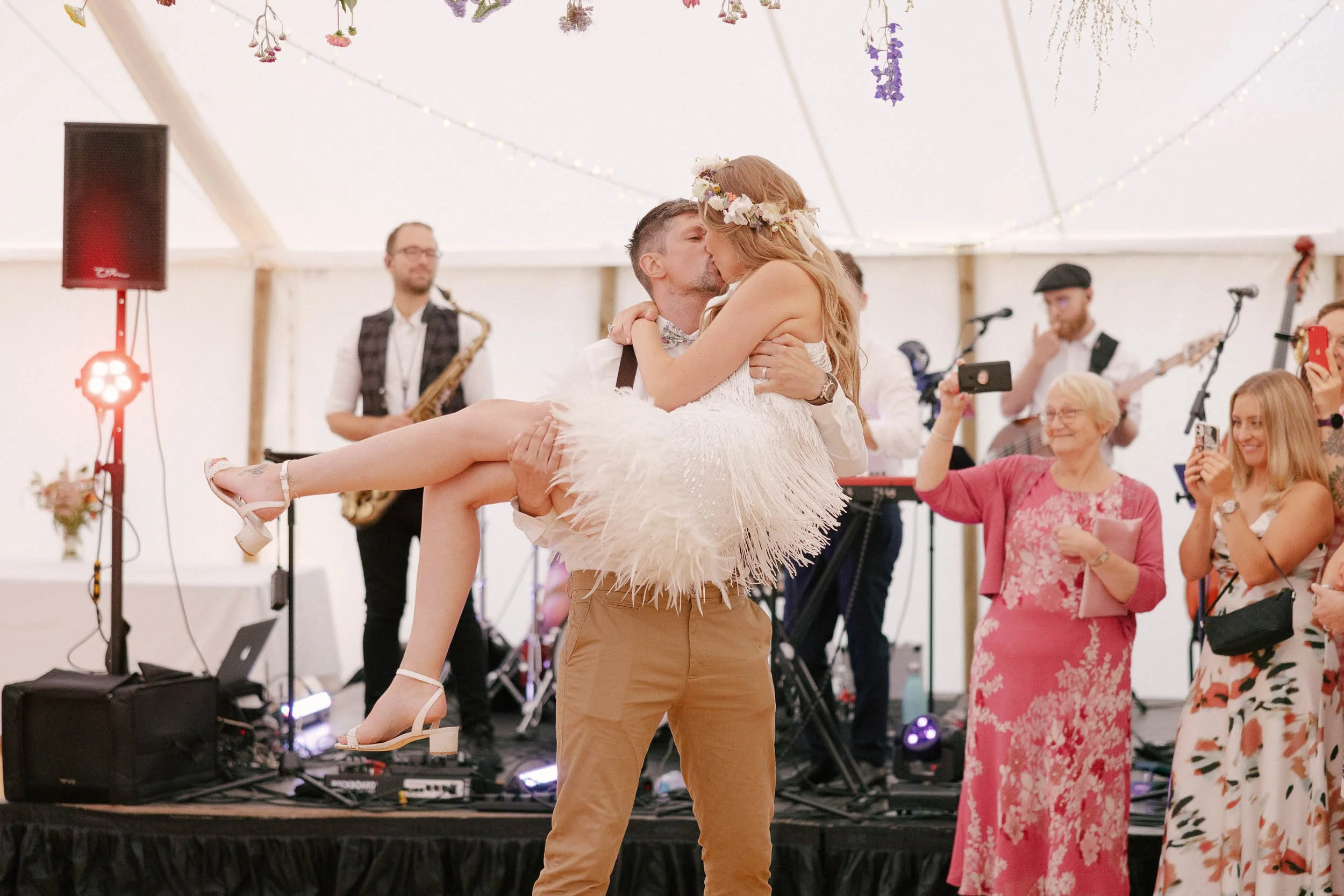 A couple sharing a first dance at their wedding reception, with the groom lifting the bride in his arms. The bride is wearing a white dress with a feathered skirt and a floral crown, while the groom is dressed in casual beige pants and a white shirt. A band and guests are visible in the background, with some guests taking photos or videos.
