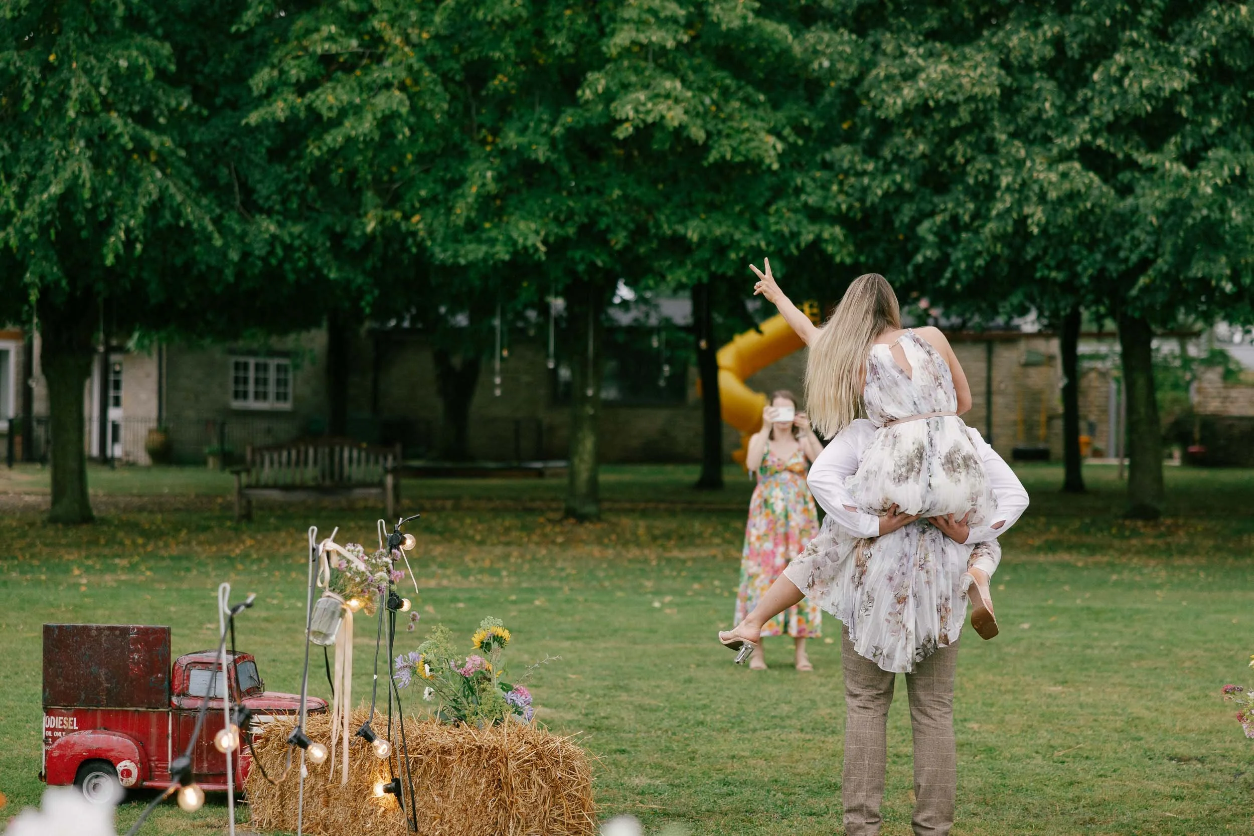 Woman with long blonde hair wearing a light-colored floral dress, sitting on the shoulders of a man in a plaid shirt and pants, making a peace sign with her hand, at an outdoor gathering with hay bales, flower arrangements, and a playground in the background.