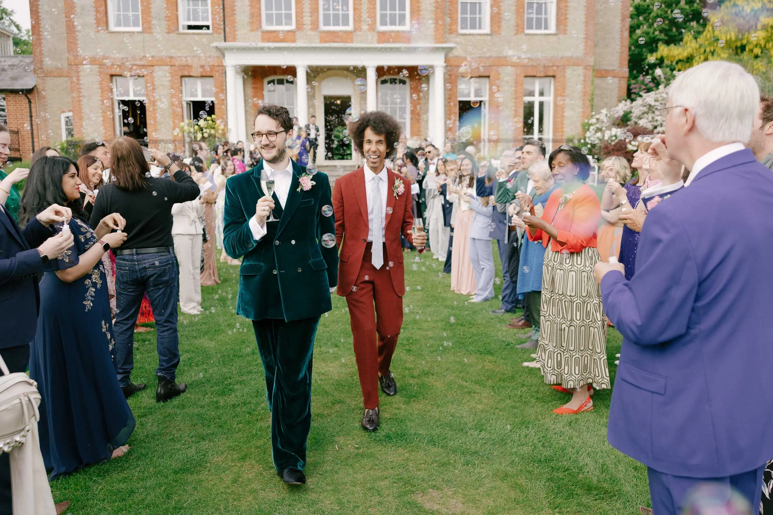 Two men in colorful suits walking through a crowd of wedding guests outside a brick building, celebrating with bubbles.