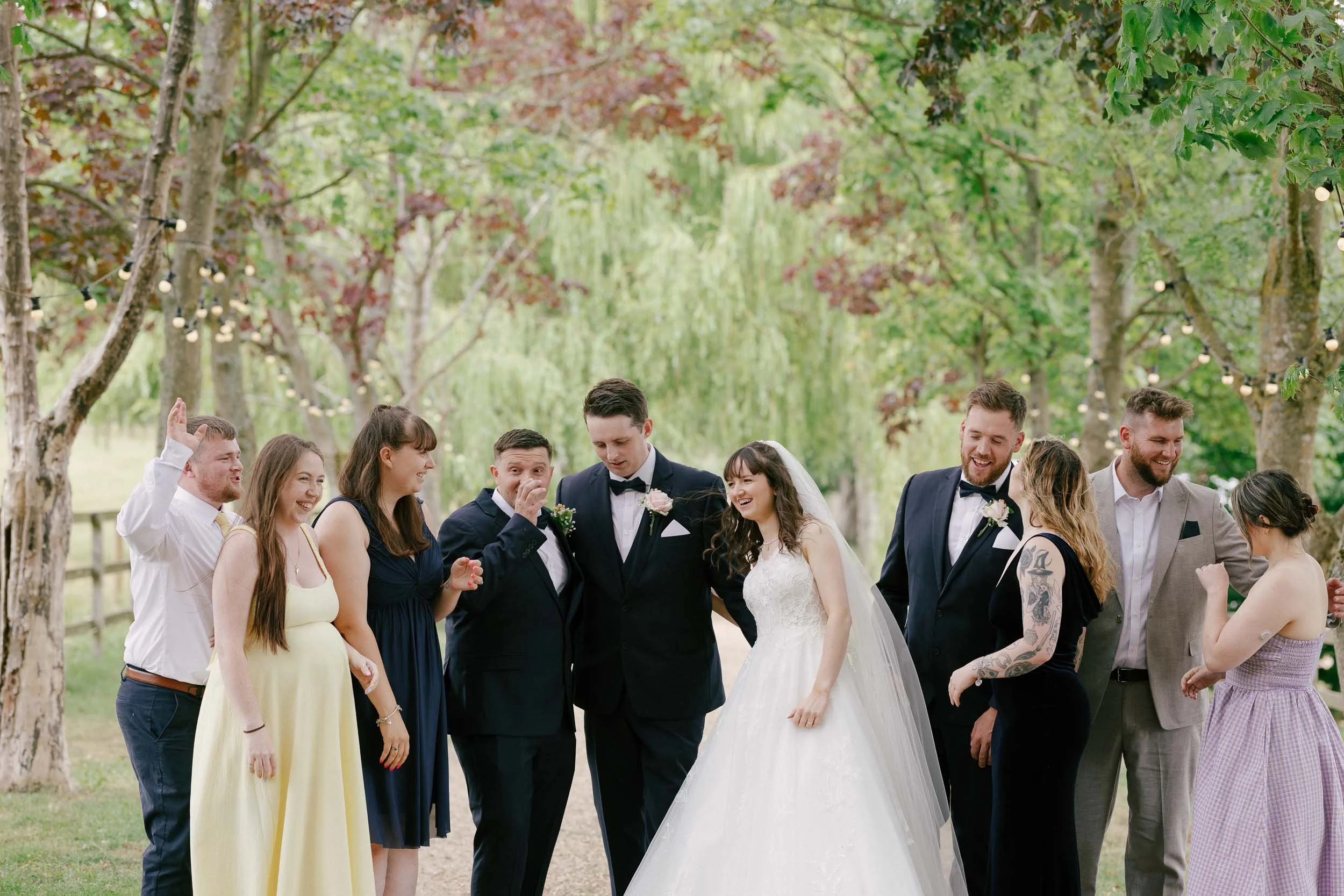 Group of people at a wedding outdoors, including bride in white wedding gown and groom in tuxedo, with friends and family, under trees with string lights.