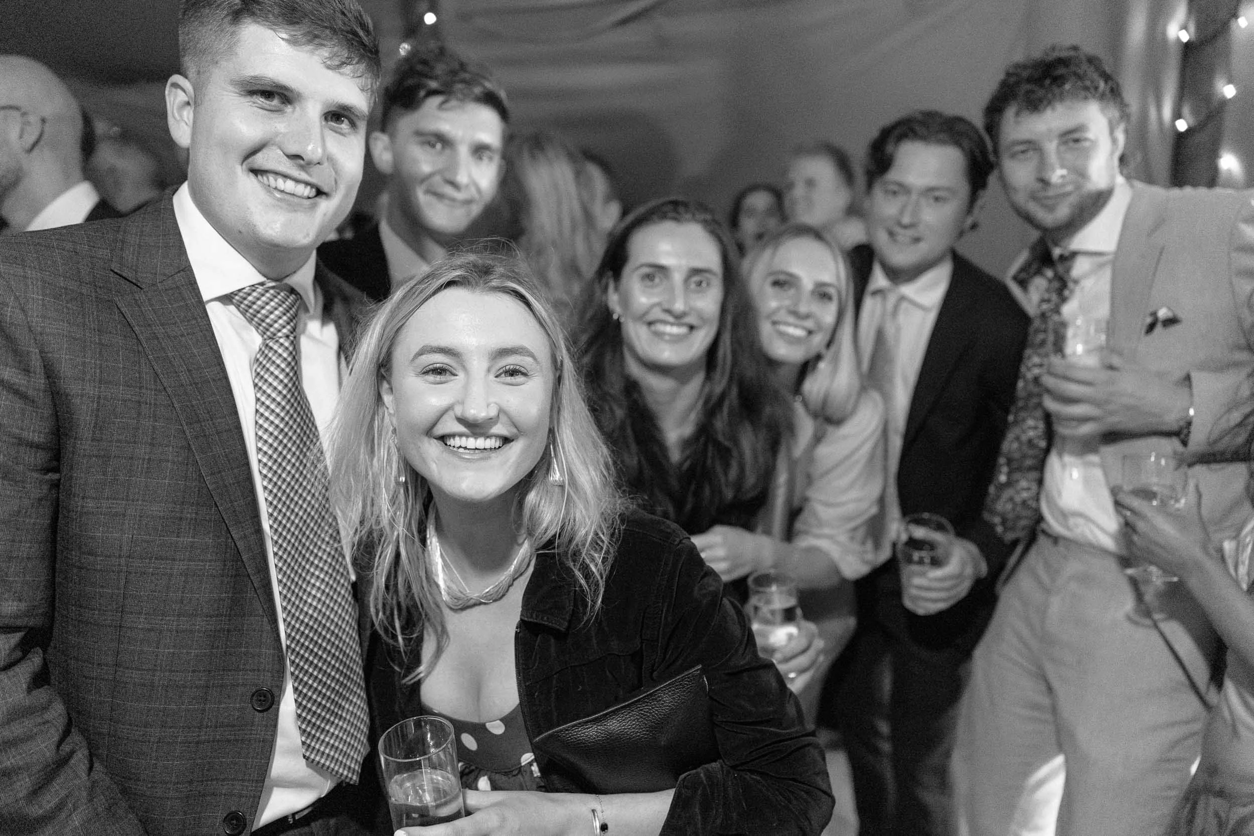 Group of seven people smiling at a party, holding drinks, dressed in formal and semi-formal attire, with string lights in the background.