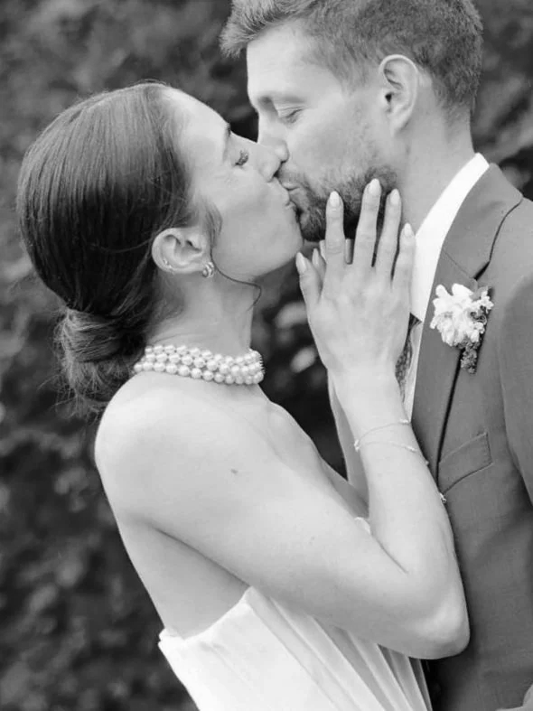 A black and white photo of a couple kissing. The woman has her hair in a bun, is wearing a strapless dress and a pearl necklace, and has her hand on the man's face. The man has short hair, a beard, and is dressed in a suit with a floral boutonniere.