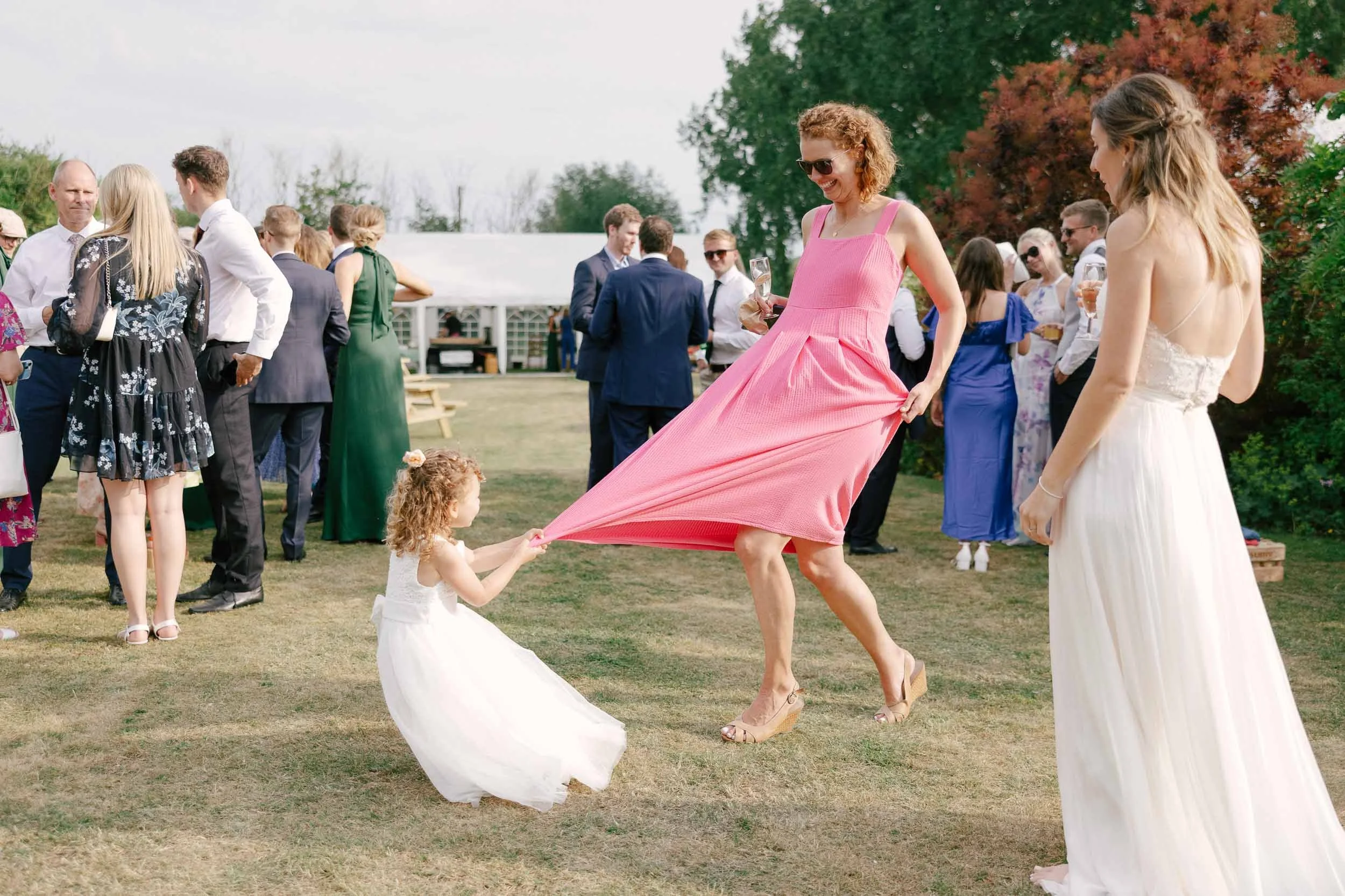 A woman in a pink dress and glasses pulling a small girl in a white dress by a pink dress at an outdoor gathering, with people socializing in the background.