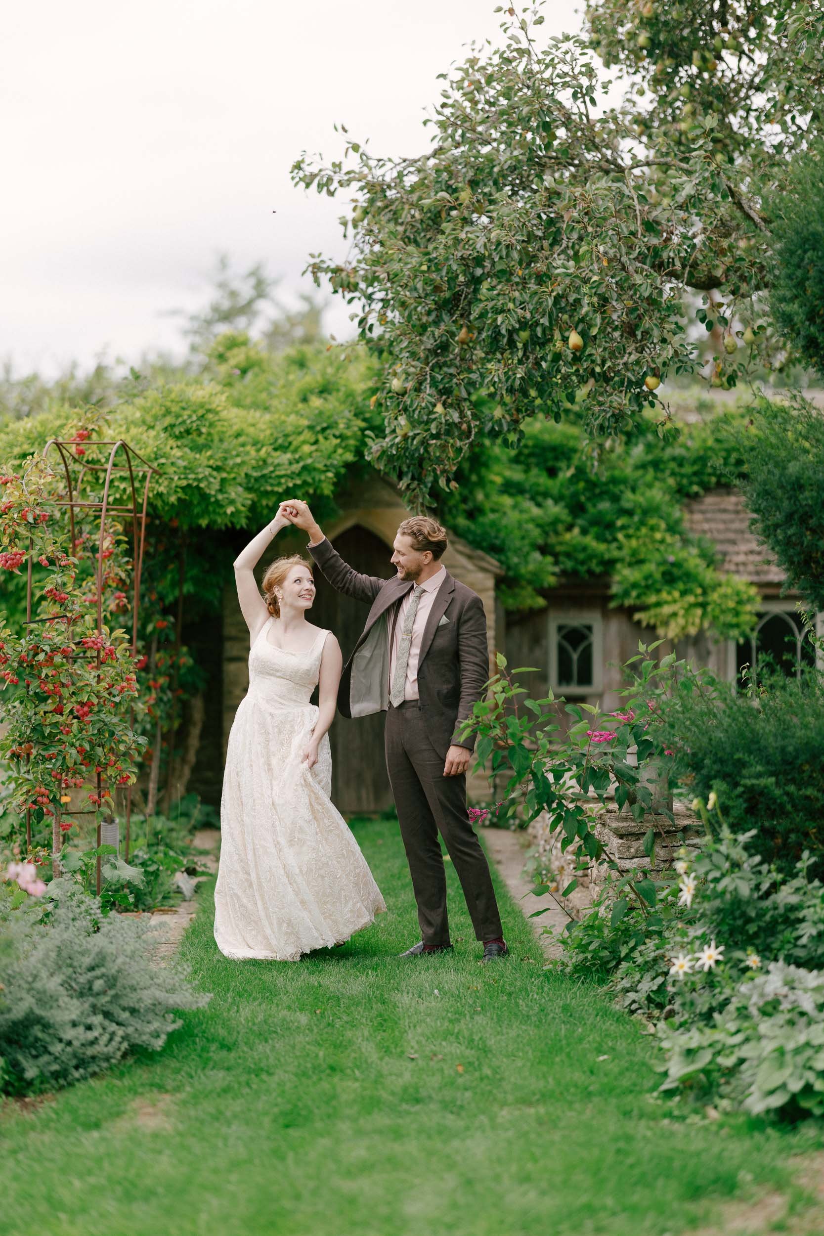 A couple dancing in a lush garden with blooming flowers and trees, dressed in vintage wedding attire.