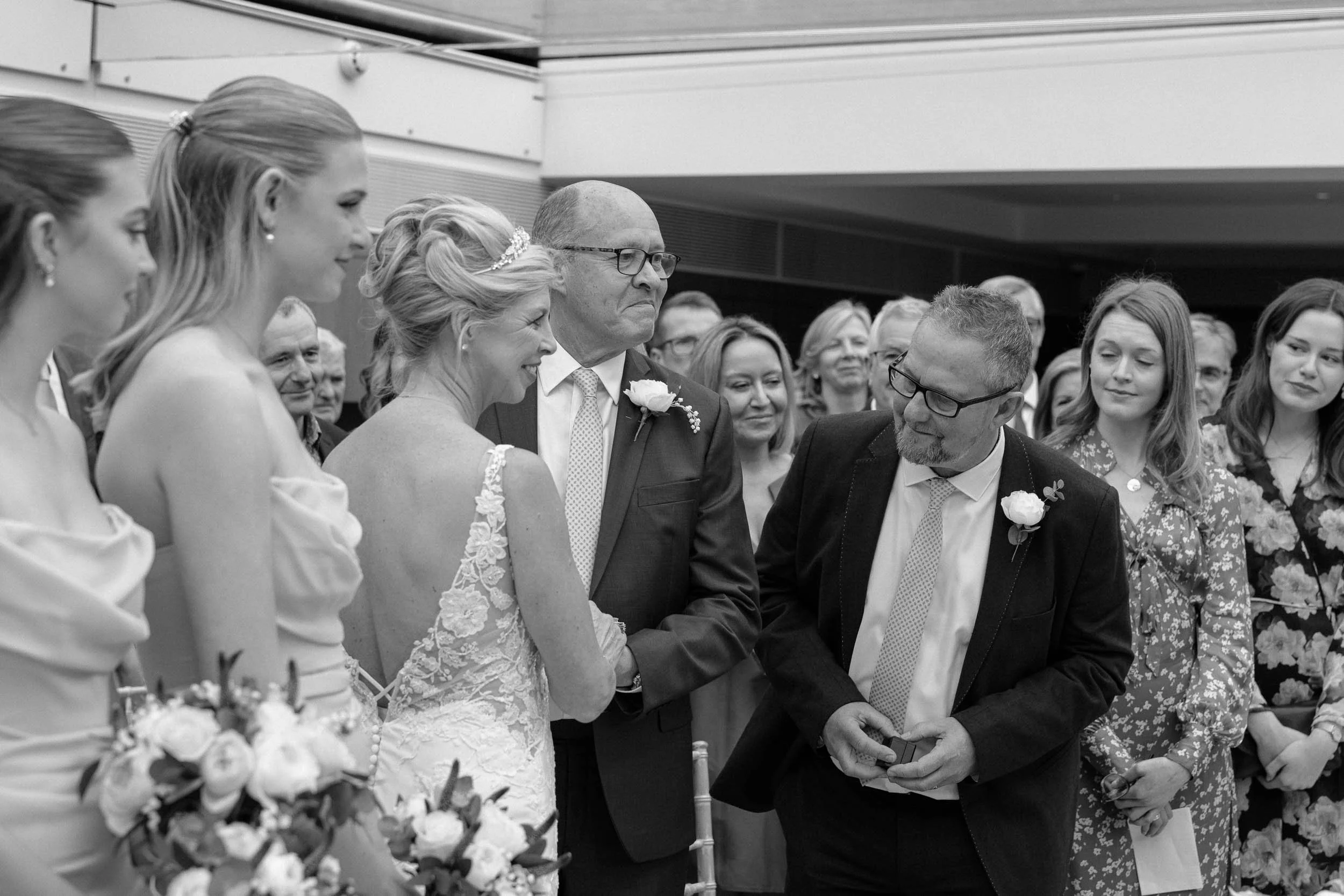 A wedding ceremony with a bride, groom, and groom's mother, surrounded by guests in an indoor venue.