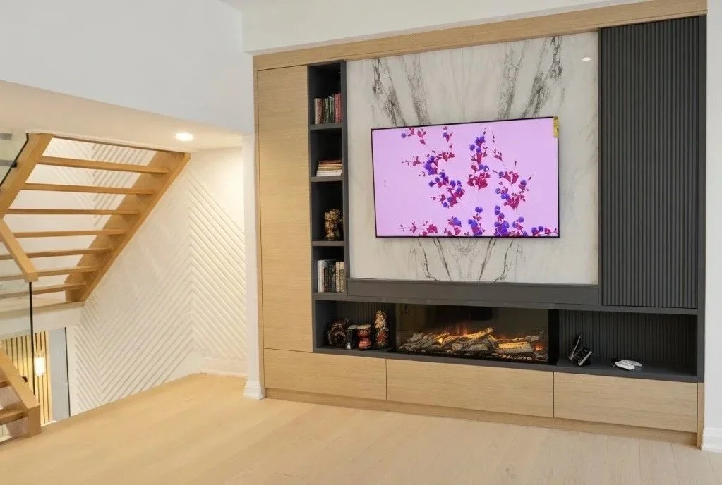 Modern living room with a wall-mounted flat-screen TV displaying a purple and pink floral pattern, a built-in fireplace beneath the TV, and wood and black paneling accents.