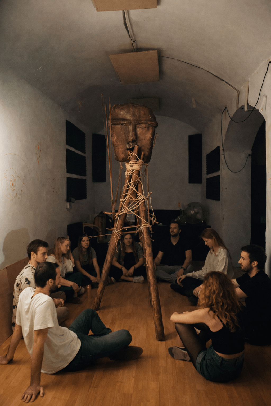 Group of people sitting in a circle in a room, observing a large wooden sculpture with a human face on top, supported by three wooden legs, with ropes tied around it.