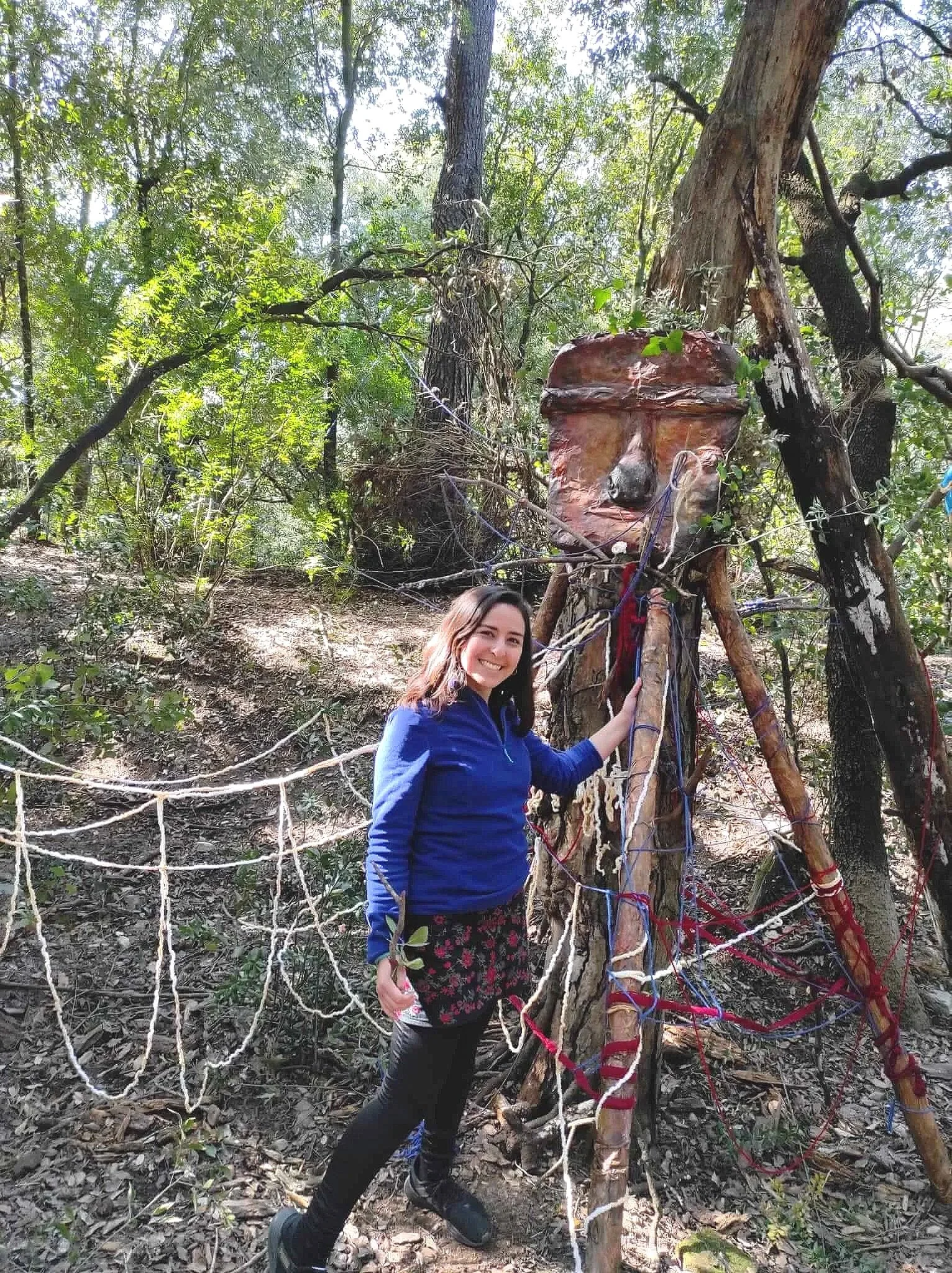 A smiling woman in a blue jacket and black leggings standing in a forest next to a wooden sculpture of a face with branches and ropes attached to it.