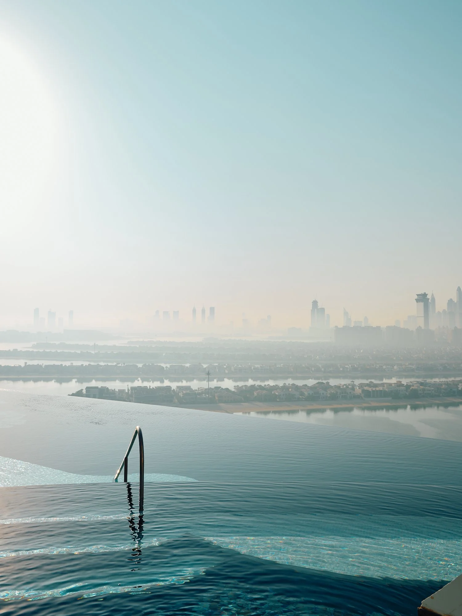 Overlooking a city skyline from an infinity pool with a metal ladder, with hazy skyscrapers in the distance under a pale blue sky.