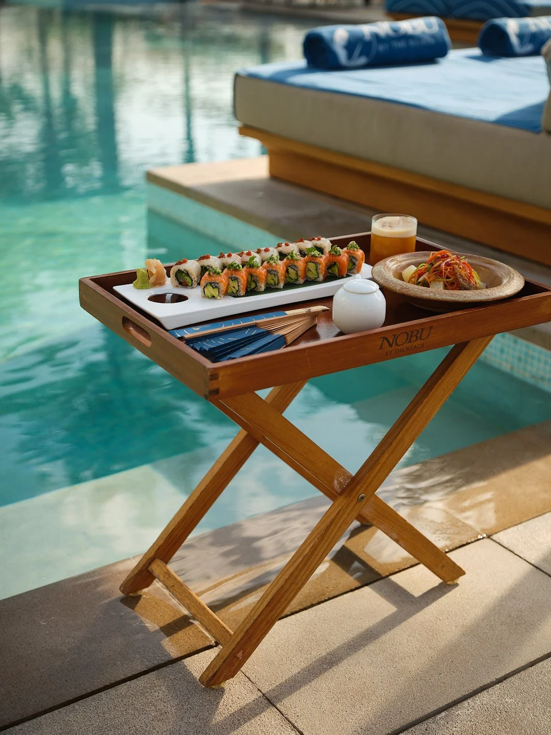 A wooden tray with sushi rolls, a small white dish, a piece of wasabi, a plate with noodles, a glass of beer, and a pair of chopsticks on a poolside area.