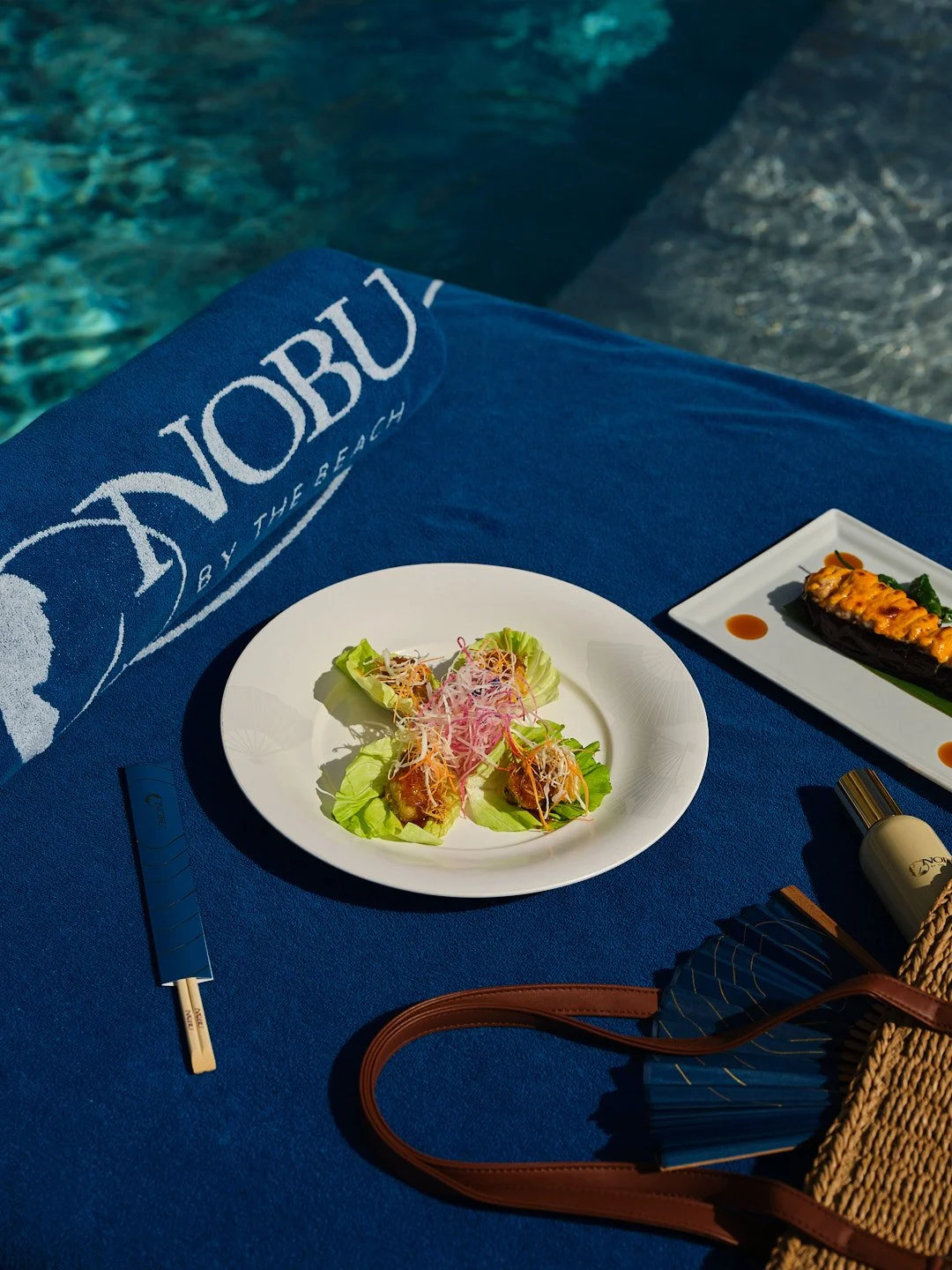 A plate of fresh salad with lettuce, shredded carrots, and other vegetables, next to a rectangular plate with cooked eggplant topped with melted cheese and sauce, both set on a blue tablecloth by a swimming pool.