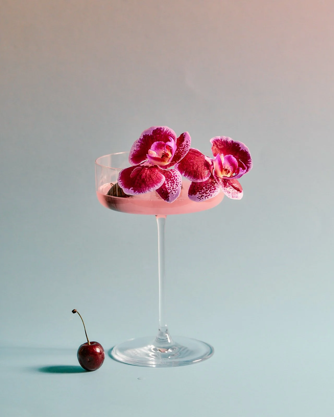 A pink cocktail in a martini glass, garnished with pink and purple orchids, with a cherry on the light blue surface beside the glass.