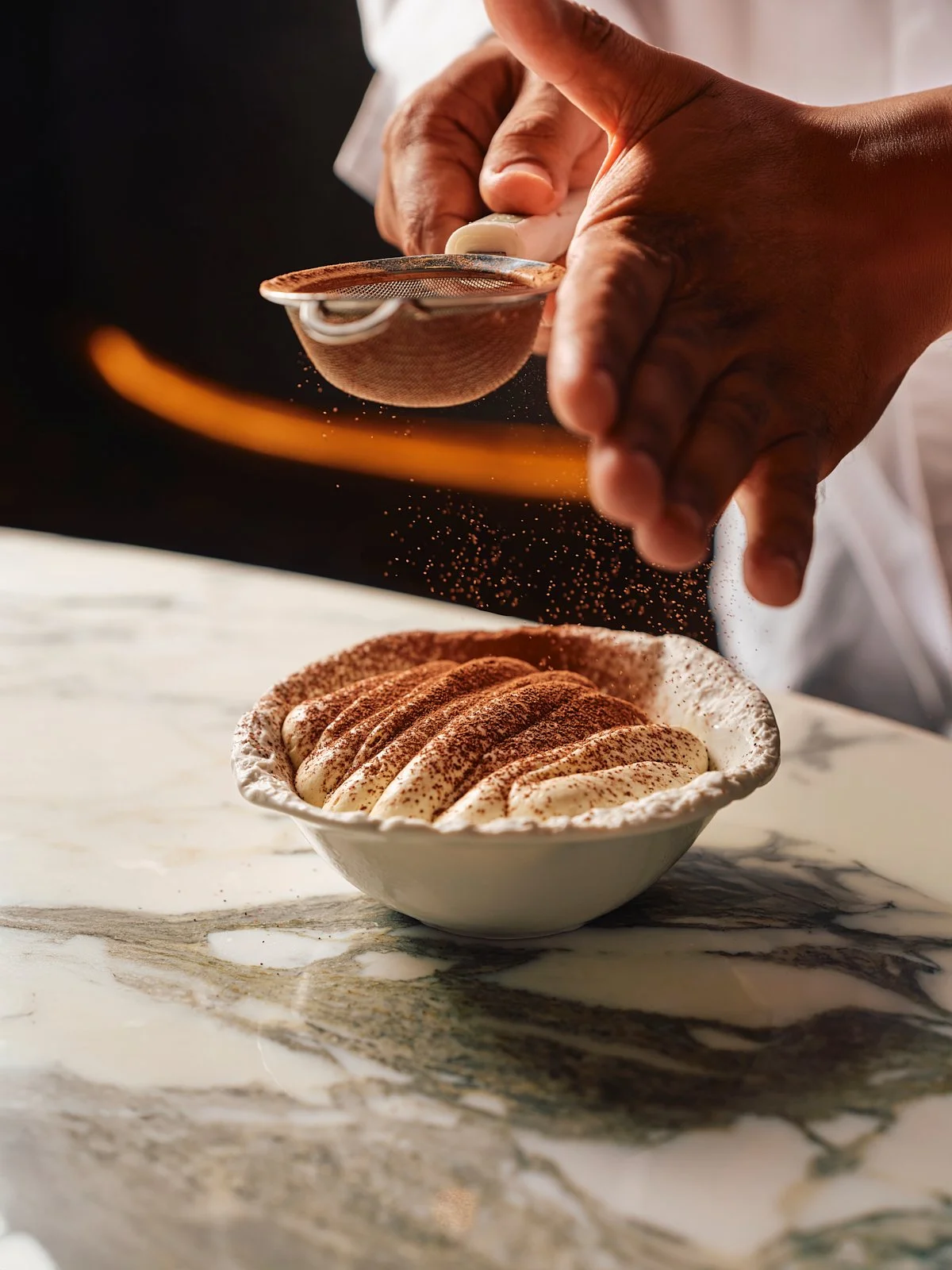 A person dusting cocoa powder onto a bowl of tiramisu on a marble surface.