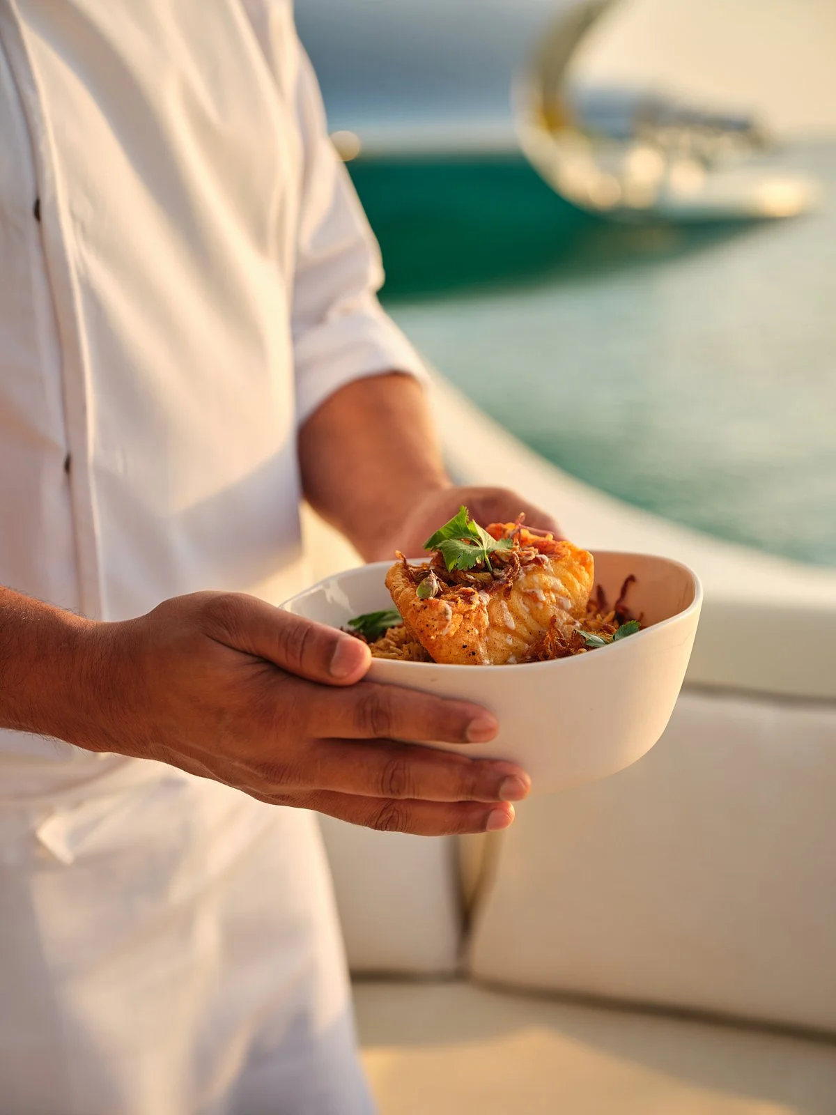 Person in white shirt holding a bowl of seafood dish garnished with herbs, with a boat and water in the background during sunset.