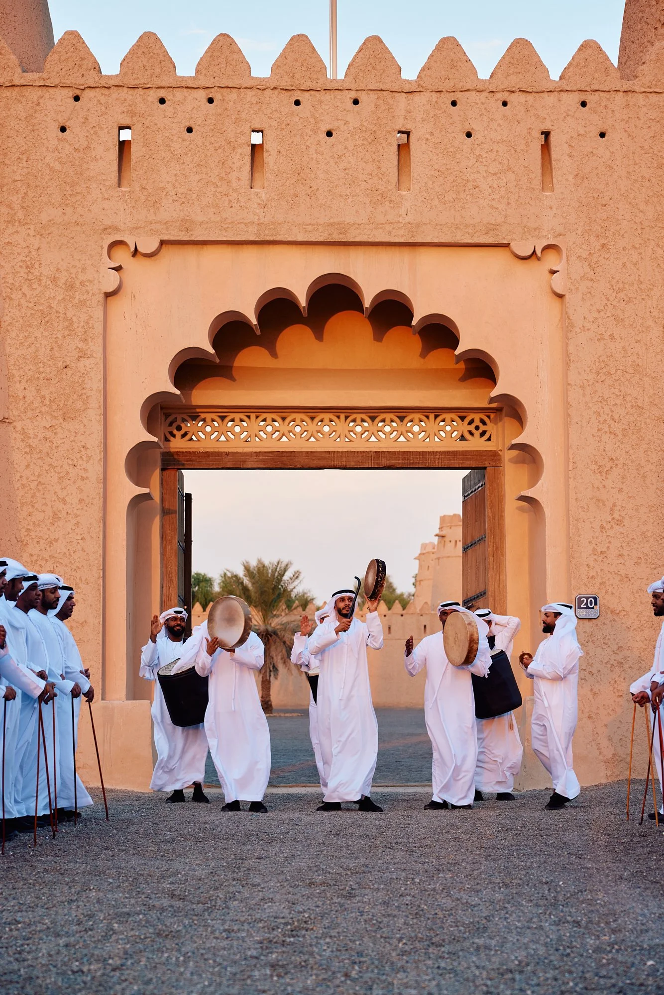 Traditional Middle Eastern dance performance with men in white robes playing drums, outside a desert fortress-style structure with an arched gateway.