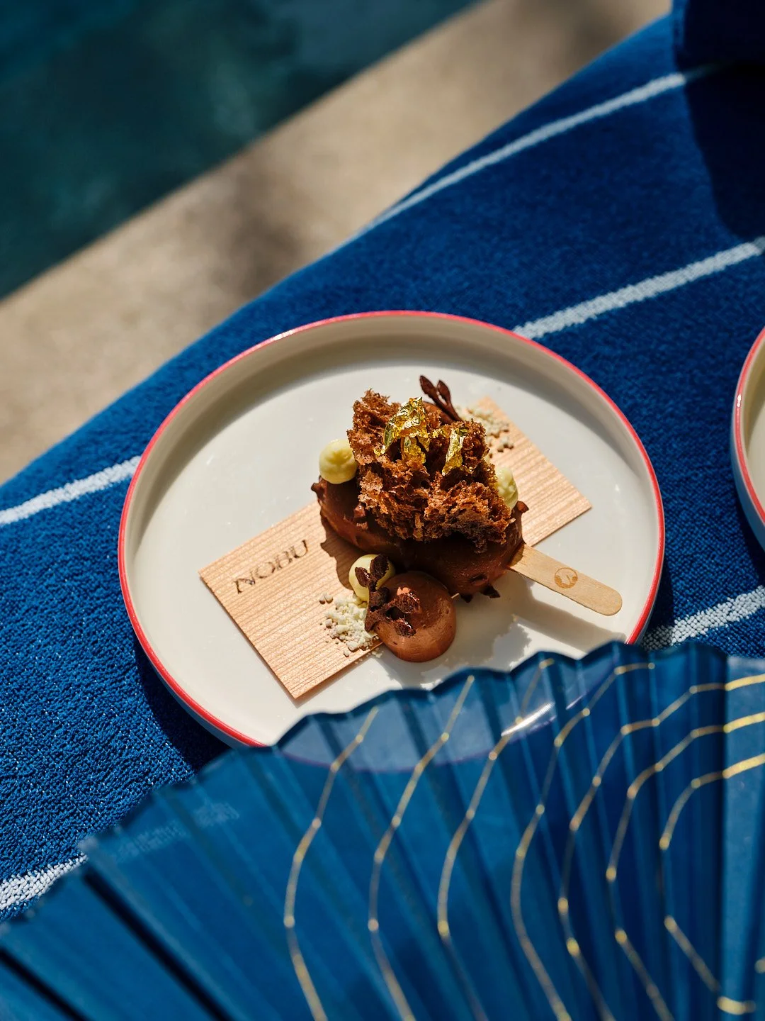 Close-up of an artisanal chocolate popsicle topped with chocolate crumble and gold leaf, served on a ceramic plate with a wooden stick and a pink rim, placed on a blue striped fabric surface.