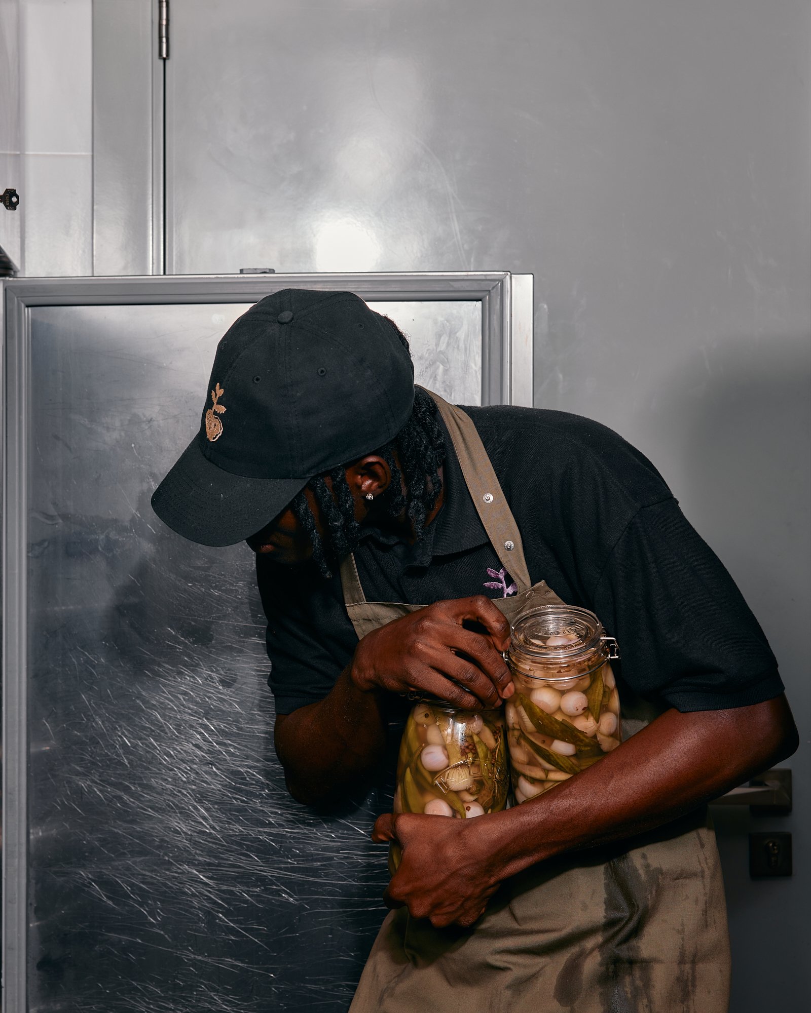 A person wearing a black cap, black shirt, and beige apron holds two glass jars filled with pickled onions and peppers.