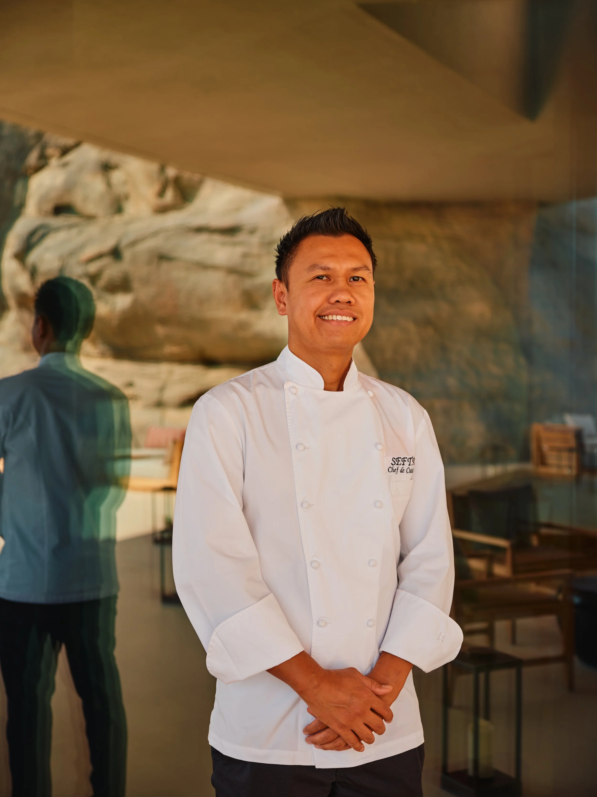 Smiling male chef in white uniform standing in a modern restaurant with a rock wall background and reflection in glass.