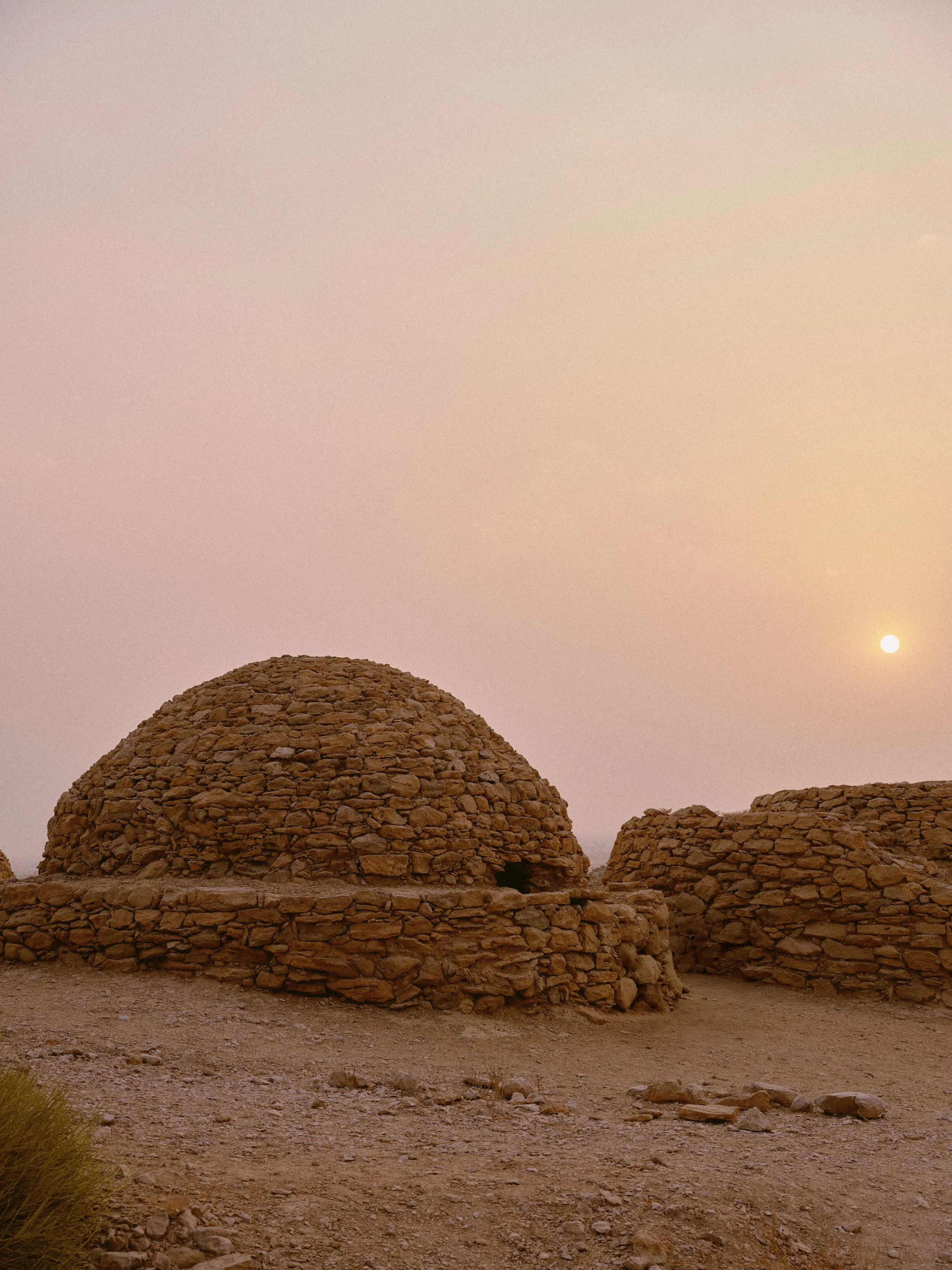 Two traditional mud and stone dome-shaped huts in an arid landscape with a setting sun and pinkish sky in the background.