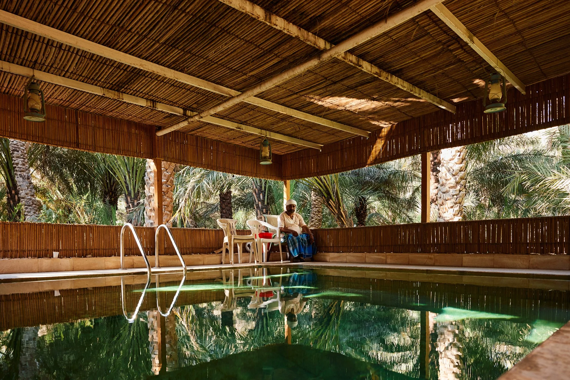 Man sitting on chair beside a swimming pool in a shaded, tropical setting with palm trees.