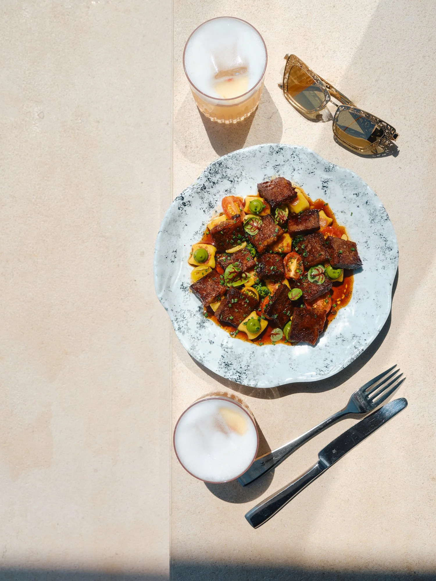 A plate of pasta with tomato sauce, topped with beef chunks and garnished with chopped herbs, on a plate with a black and white look, with two glasses of iced beverages and a pair of sunglasses nearby.